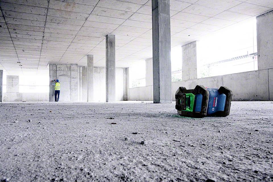 Betonbaustelle mit mehreren Säulen, ein Bauarbeiter im Hintergrund, im Vordergrund ein grünes Gerät auf dem Boden.