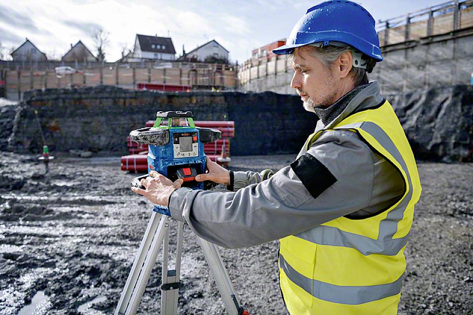 Ein Bauarbeiter mit blauem Helm und gelber Warnweste bedient ein Vermessungsgerät auf einer Baustelle mit Erdarbeiten im Hintergrund.