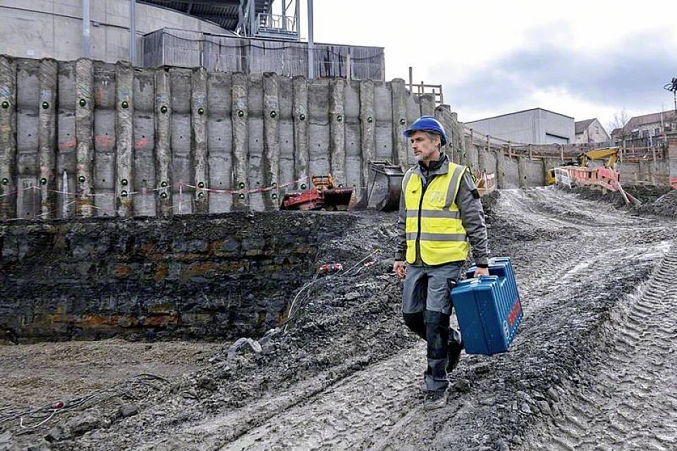 Bauarbeiter mit Helm und Warnweste geht über eine Baustelle, hält einen Werkzeugkoffer. Im Hintergrund Baugerüste und Maschinen.