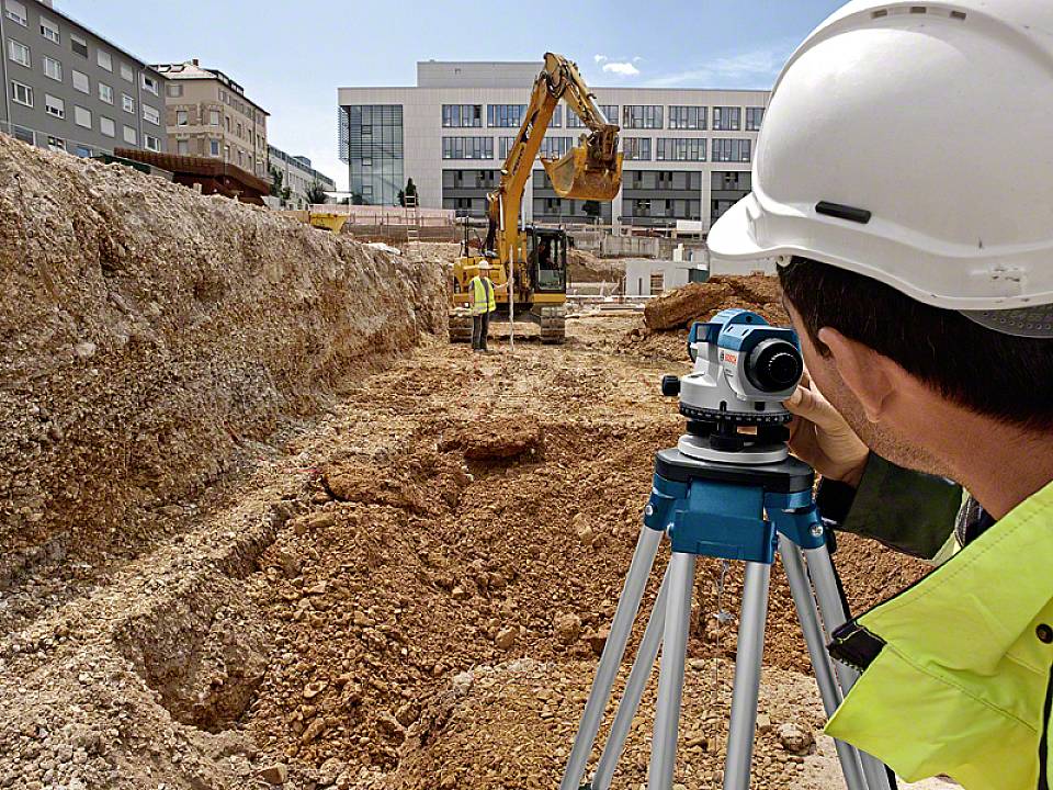 Ein Bauingenieur mit Schutzhelm benutzt ein Vermessungsgerät auf einer Baustelle, während im Hintergrund ein Bagger arbeitet.