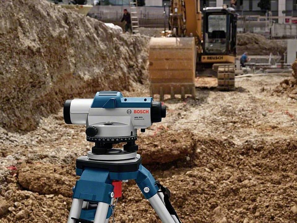 Eine Baustelle mit einem Bau-Nivelliergerät im Vordergrund und einem Bagger, der im Hintergrund Erde bewegt.