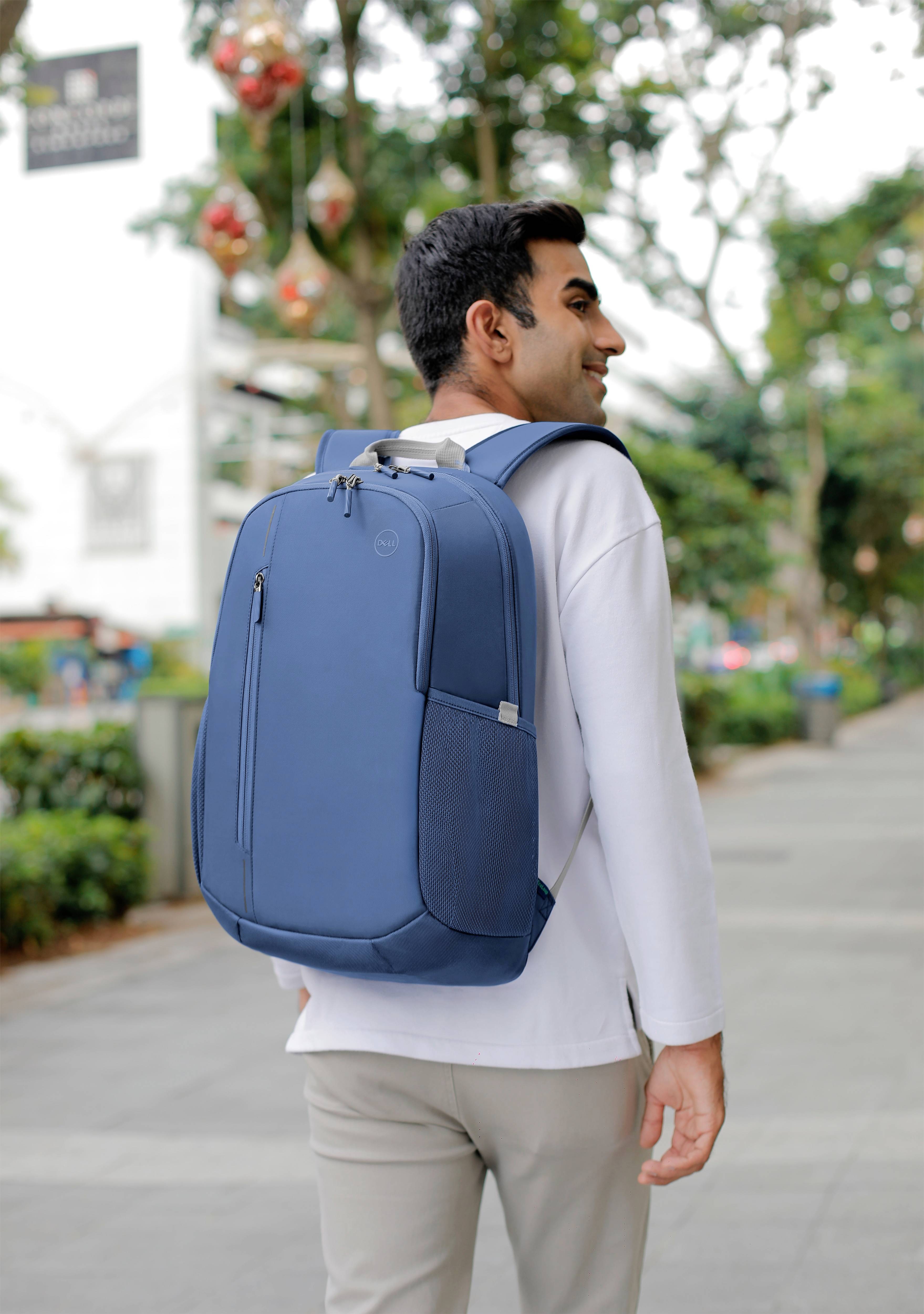 Eine Person mit einem blauen Rucksack geht lächelnd auf einem Gehweg im Freien. Im Hintergrund sind Bäume und Dekorationen.