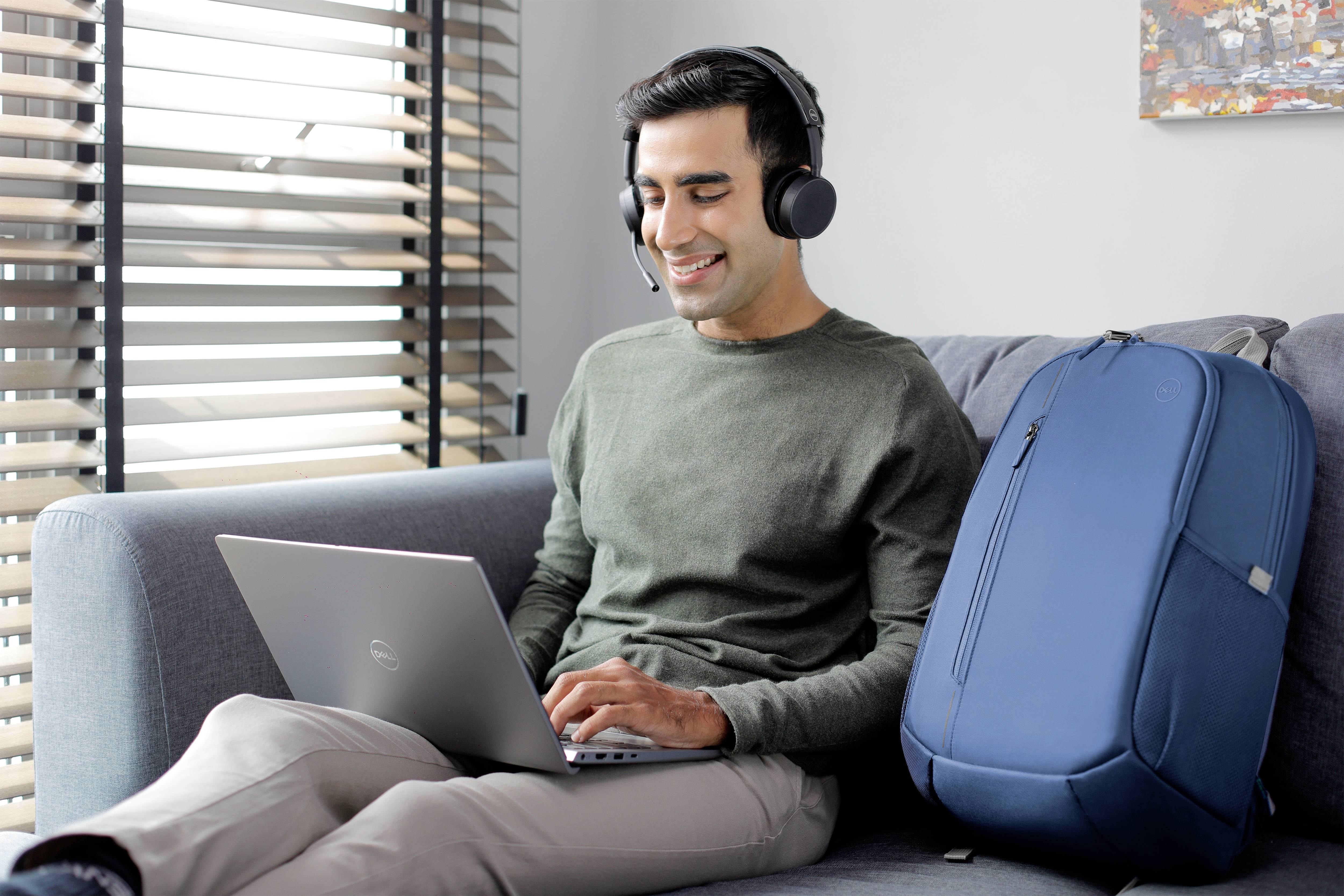 Ein Mann mit Kopfhörern sitzt lächelnd auf einem Sofa und benutzt einen Laptop. Neben ihm steht ein blauer Rucksack.
