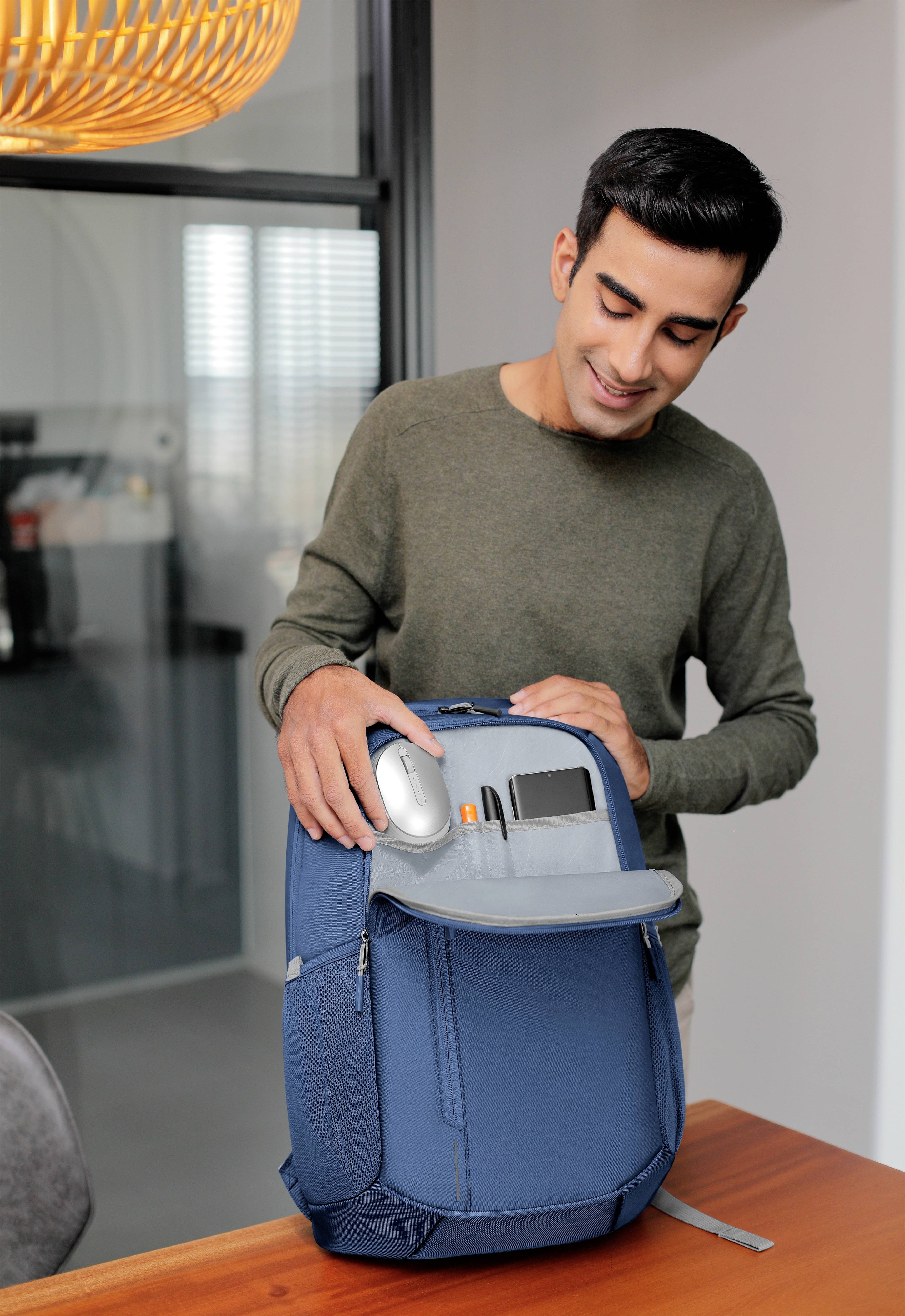 Ein Mann steht an einem Tisch und lächelt, während er die Vordertasche eines blauen Rucksacks öffnet. Im Hintergrund ist ein modern eingerichteter Raum.