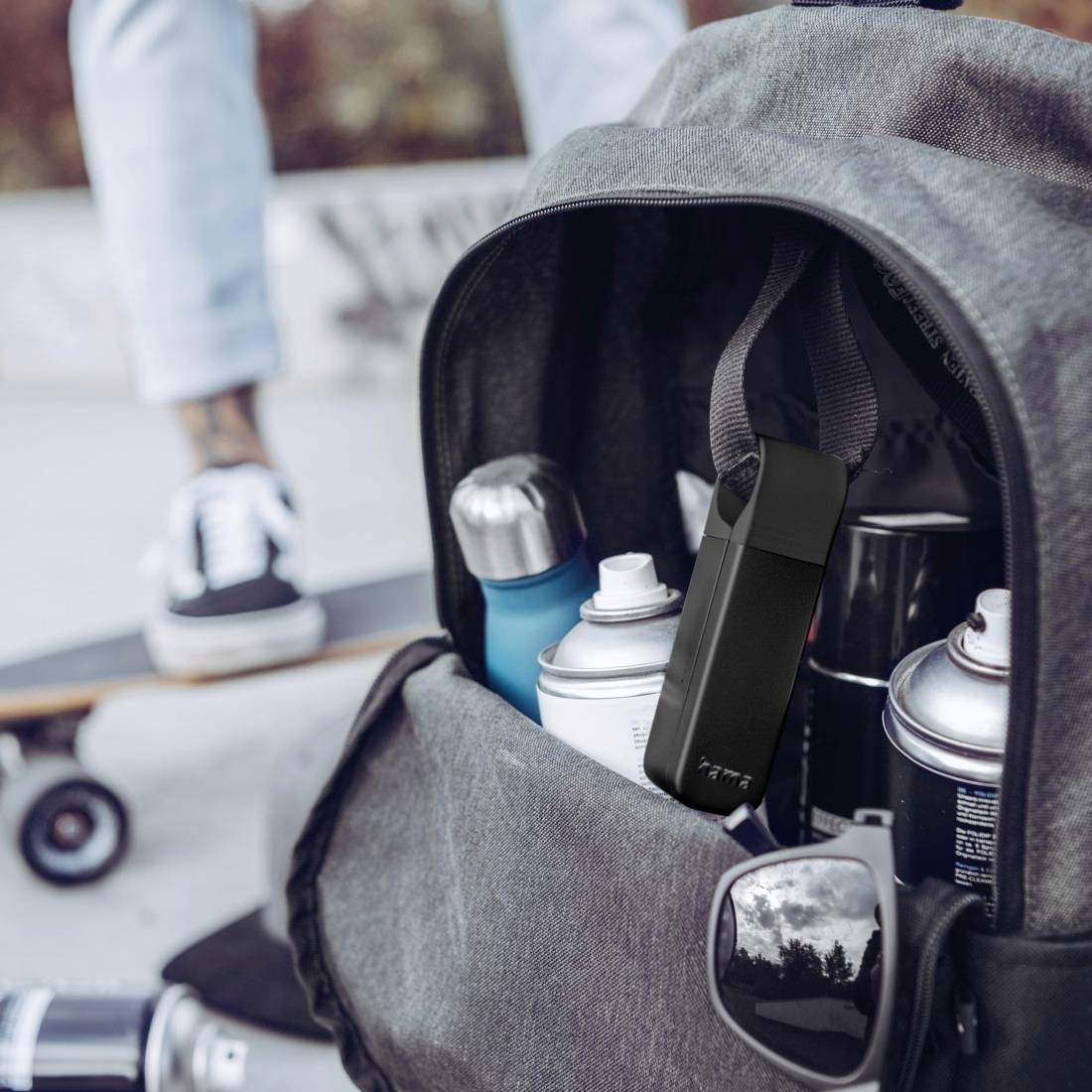 Ein Rucksack gefüllt mit Spraydosen, einer Wasserflasche und Sonnenbrille; Im Hintergrund unscharf ein Skateboard und eine Person mit Turnschuhen.