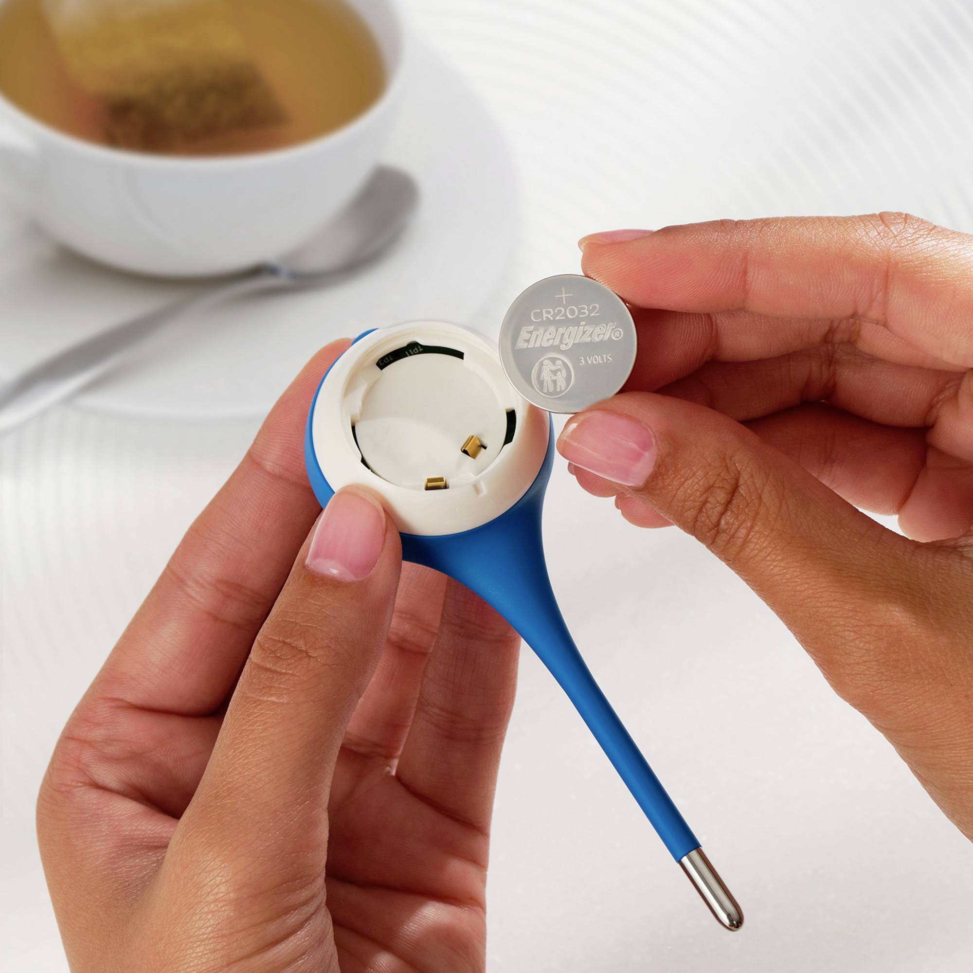 Eine Person hält eine Knopfzellenbatterie der Marke 'Energizer CR2032' neben einem blauen Gerätegehäuse auf einem Tisch.