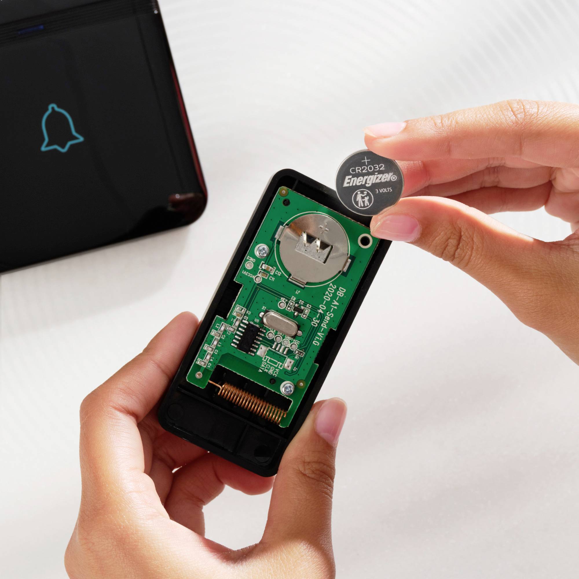 Eine Hand ersetzt eine Knopfzellenbatterie in einem kleinen elektronischen Gerät, während ein anderes Gerät mit Glockensymbol daneben liegt.