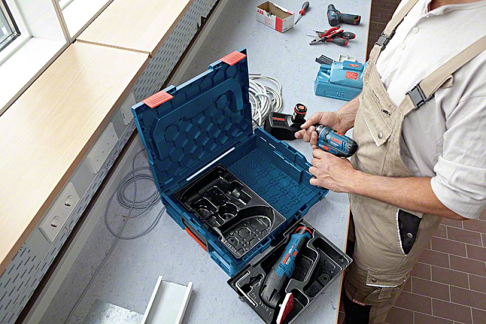 Eine Person in Arbeitskleidung hält ein Elektrowerkzeug und bereitet es für den Gebrauch vor. Auf dem Tisch liegen verschiedene Werkzeuge und Kabel.