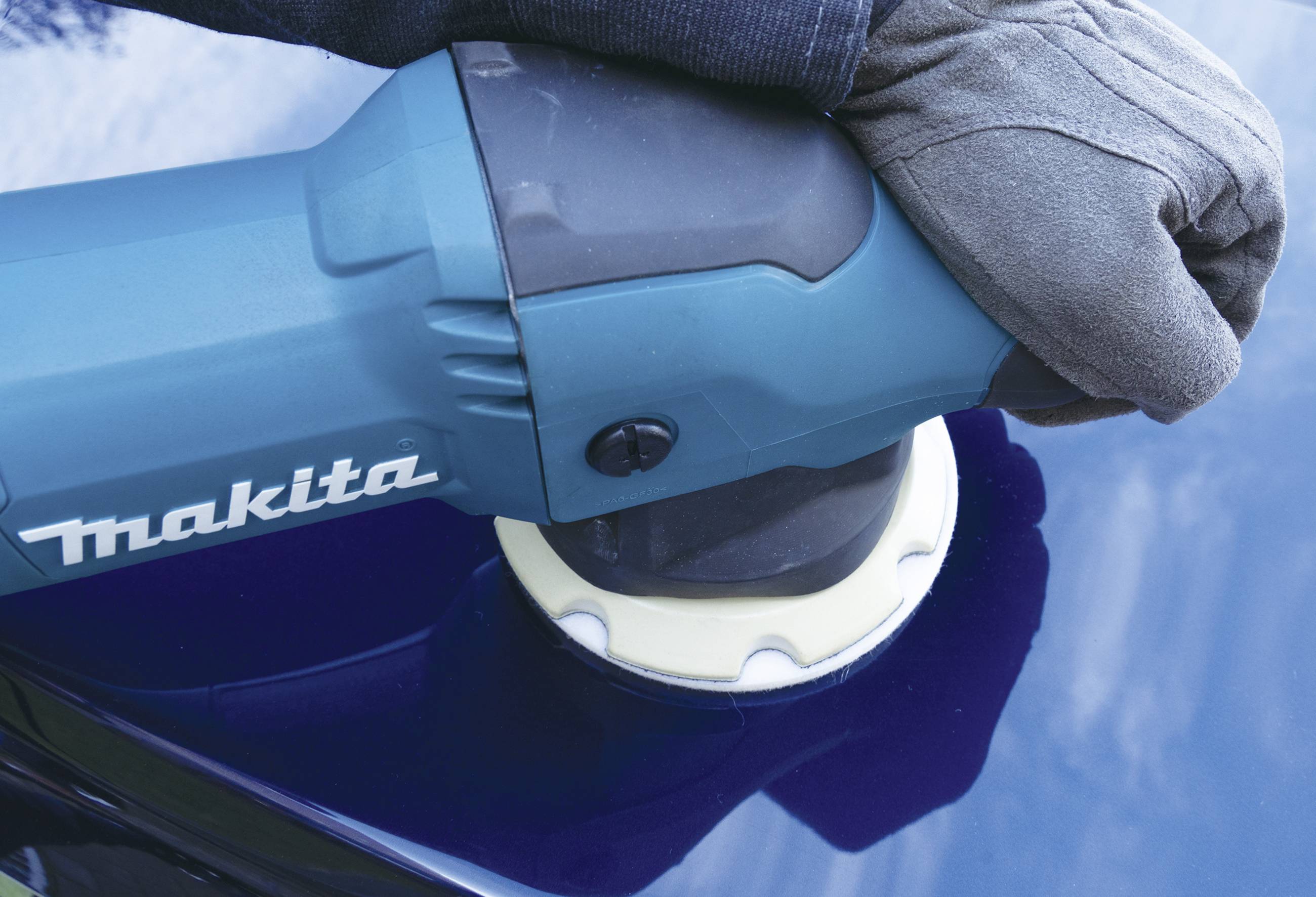 Eine Hand mit Handschuh hält eine Makita-Poliermaschine, die eine blaue Oberfläche poliert.