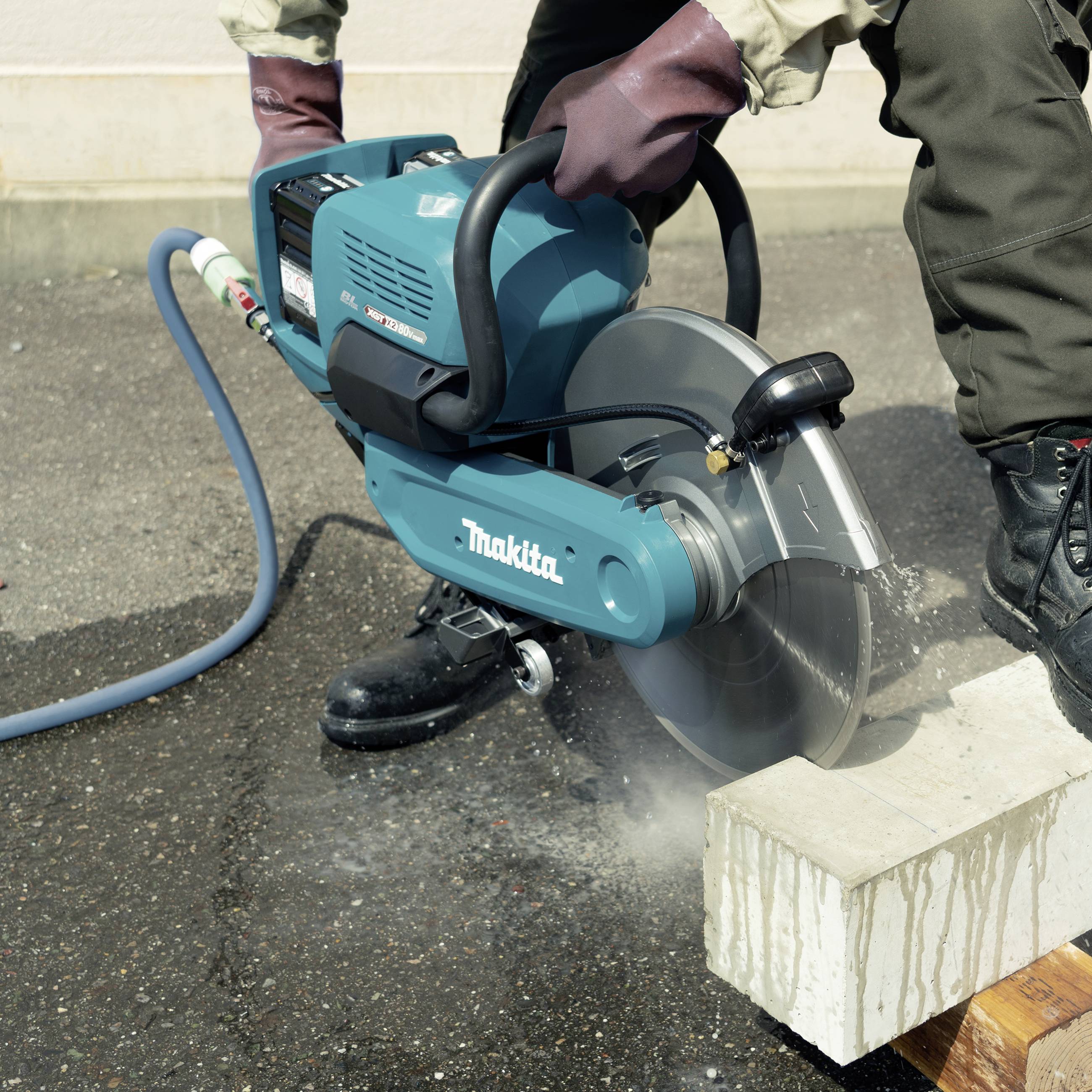 Person schneidet Betonstein mit einer motorisierten Säge auf Asphalt, trägt Handschuhe und Schutzkleidung.