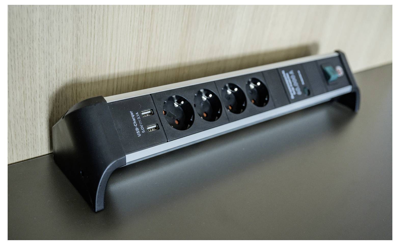 Schwarze Steckdosenleiste mit vier Steckdosen und zwei USB-Anschlüssen, die an einer Wand auf einem Schreibtisch oder Regal montiert ist.