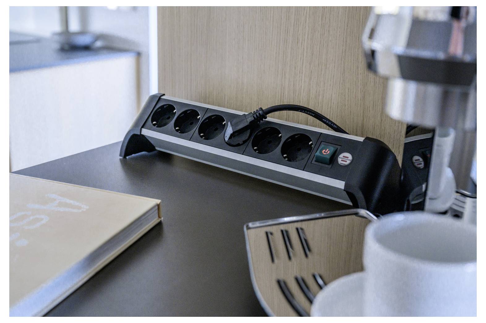 Steckerleiste mit sechs Steckdosen und angeschlossenem Kabel auf einem Schreibtisch neben einer Kaffeemaschine und einer Tasse, was einen Arbeitsbereich oder eine Pausenzone andeutet.