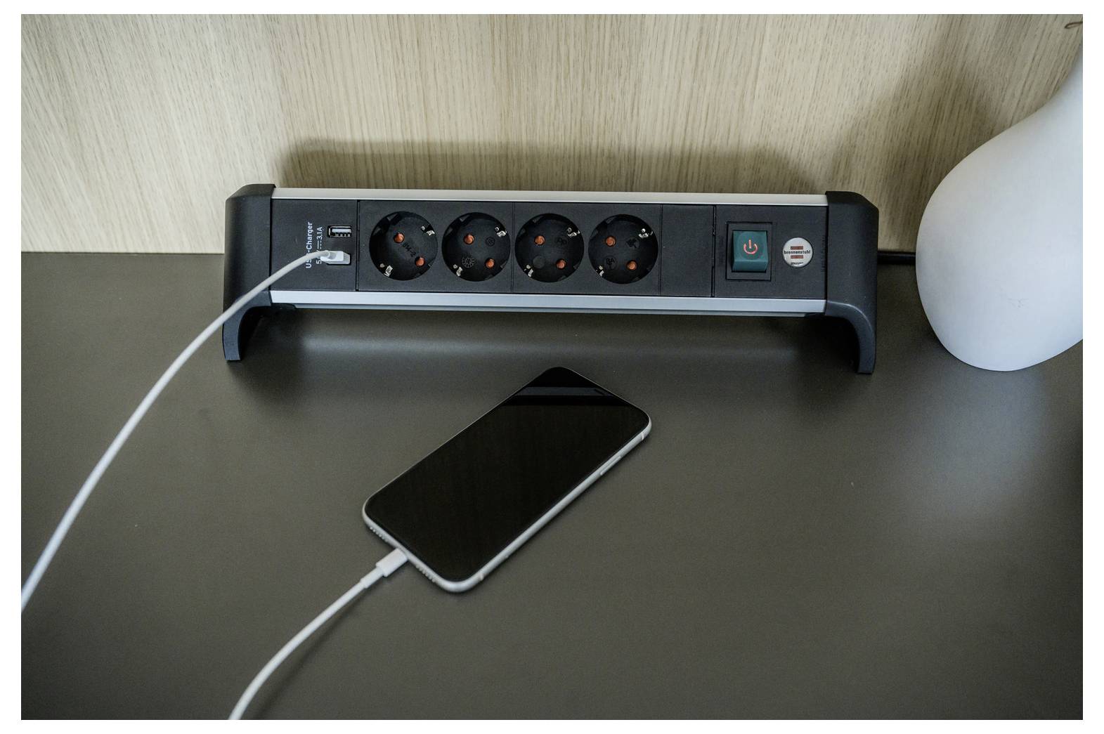 Ein Smartphone lädt auf einem Schreibtisch neben einer Steckdosenleiste mit mehreren Steckplätzen und einer Lampe.