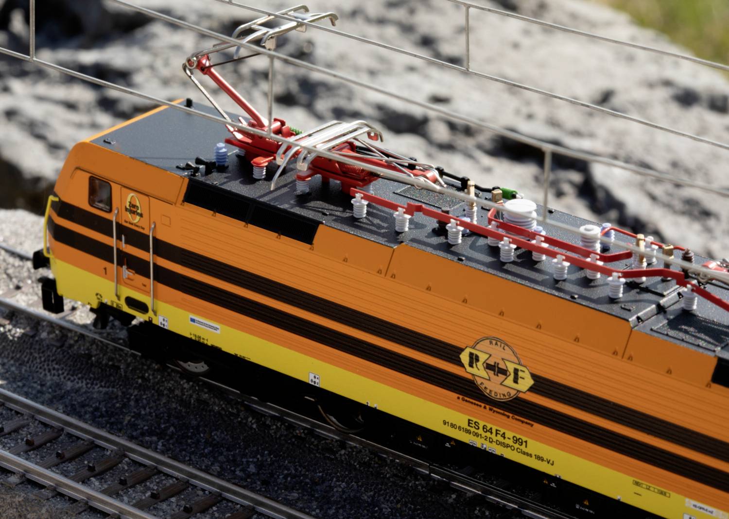Spielzeugmodell einer gelb-schwarzen Elektrolok mit Detailansicht der Oberleitung, auf Schienen vor felsigem Hintergrund platziert.