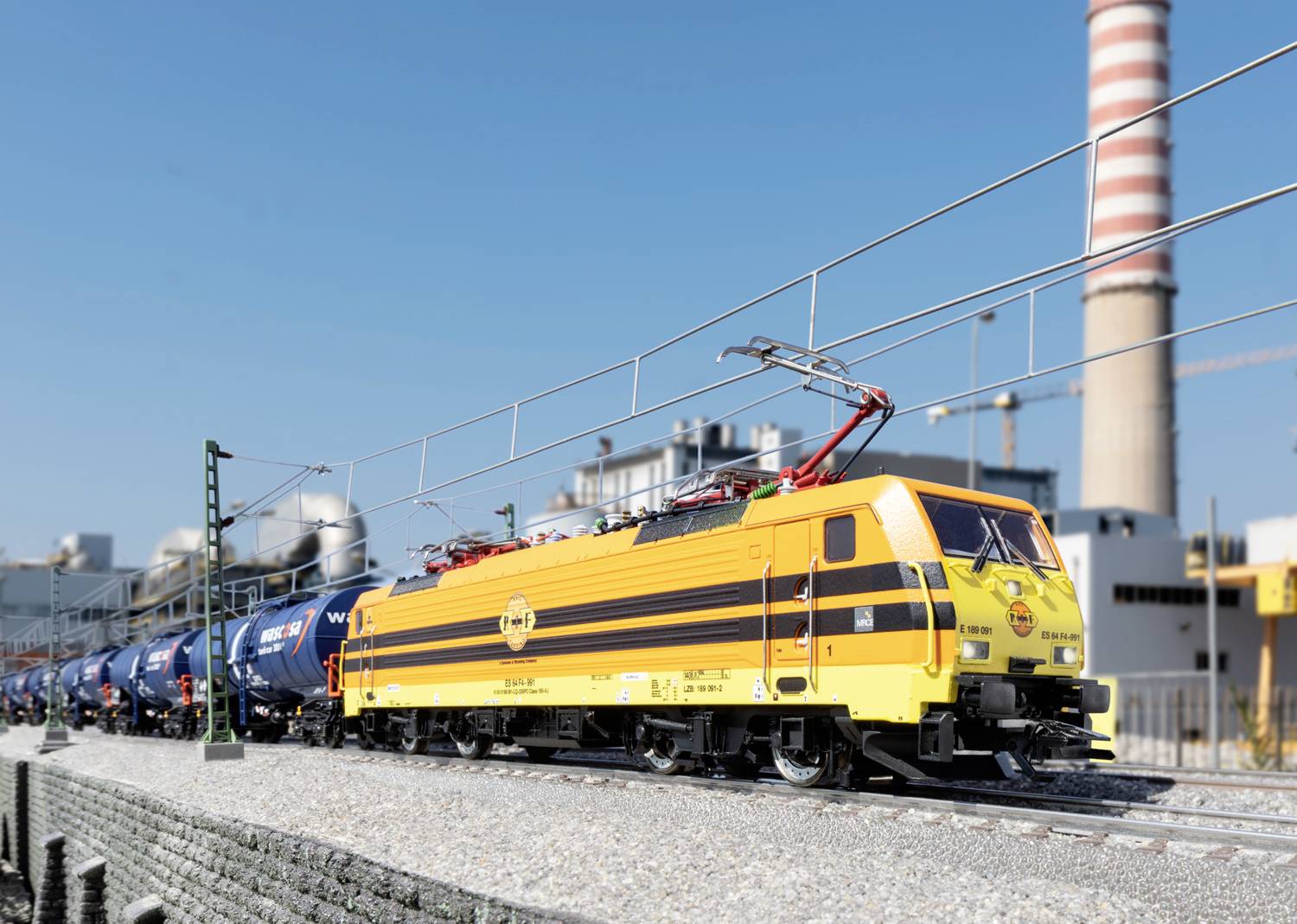 Gelbe Lokomotive zieht Güterzug mit blauen Kesselwaggons entlang Industrieanlage, im Hintergrund Schornstein unter blauem Himmel.