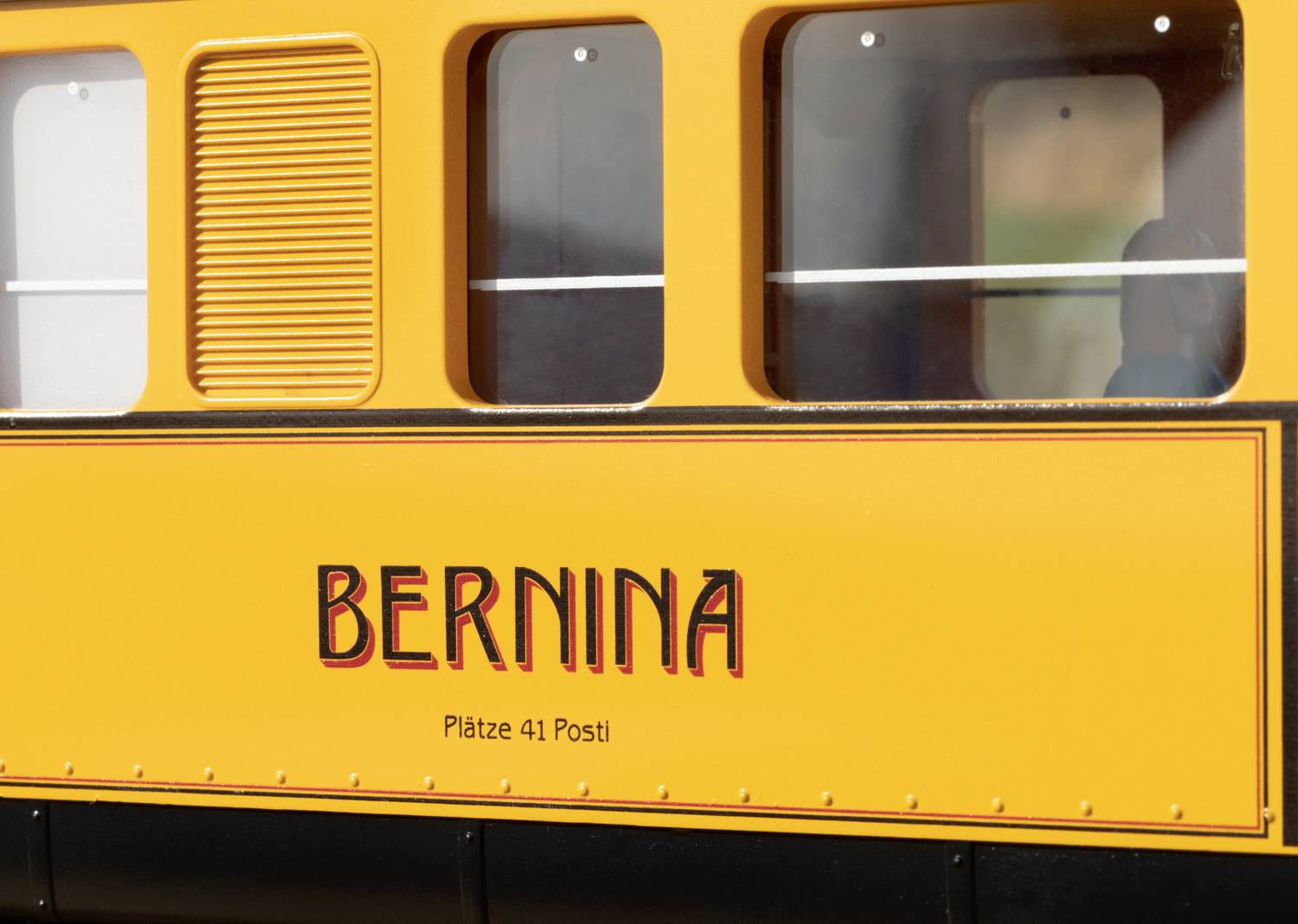 Gelbes Zugabteil mit der Aufschrift 'Bernina'. Darunter steht 'Plätze 41 Post'. Fenster und Details der Waggonwand sind sichtbar.