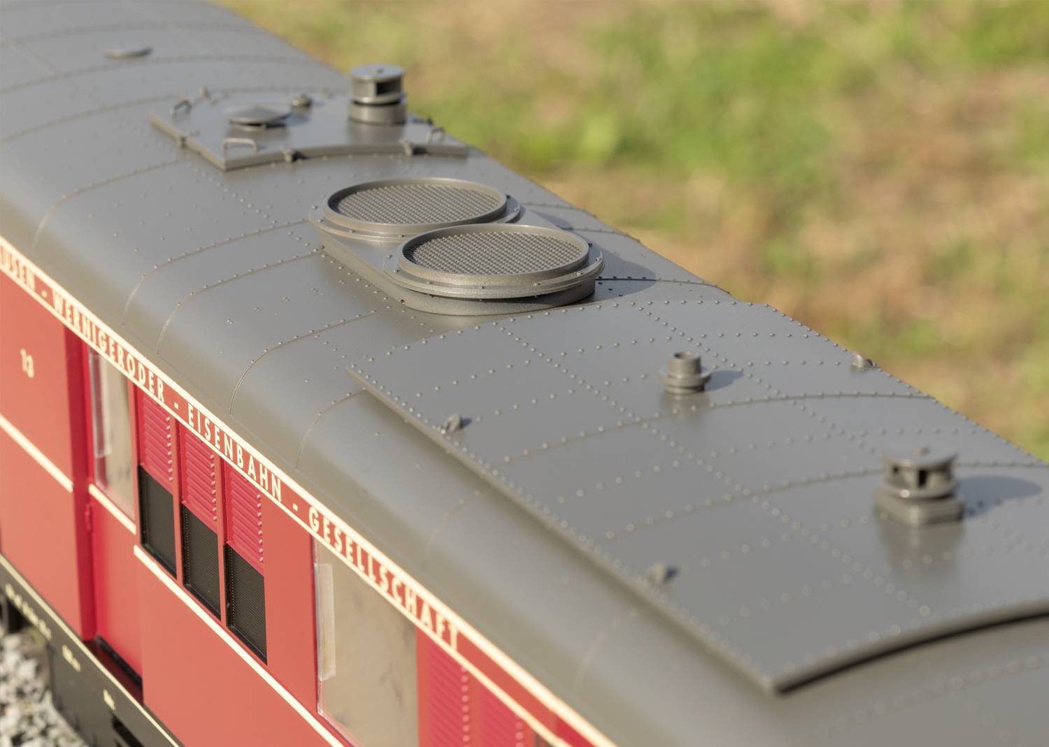 Nahaufnahme eines roten Modelleisenbahnwagens mit grauem Dach und Lüftungsschlitzen. Im Hintergrund unscharfes Gras sichtbar.