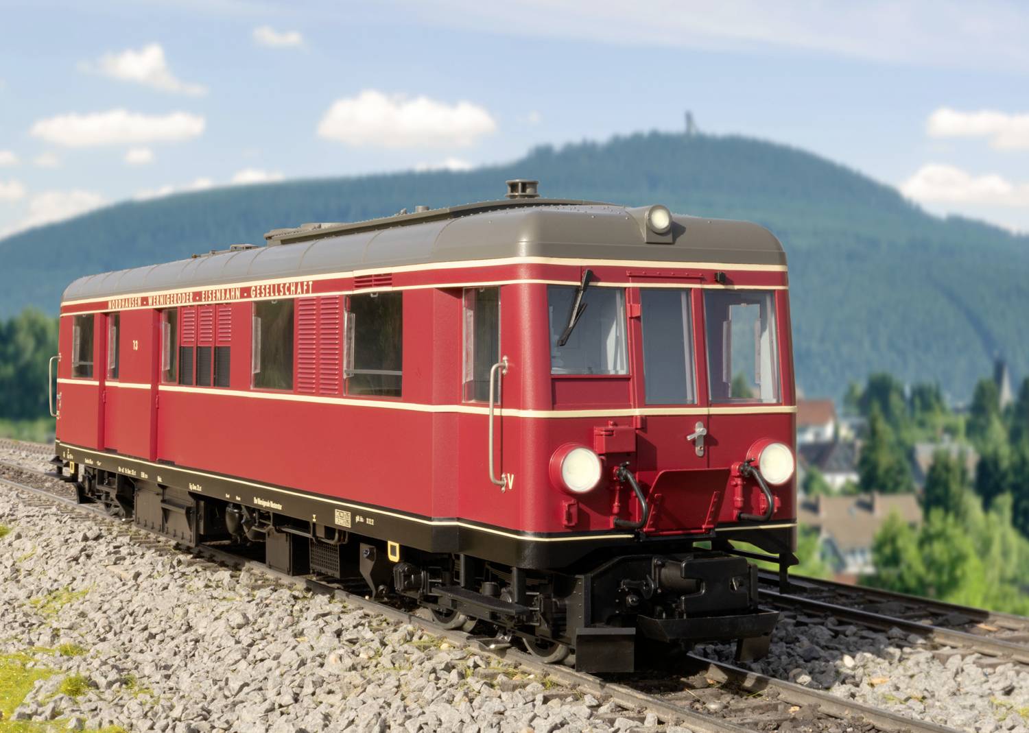 Ein roter, historischer Triebwagen fährt auf Schienen durch eine hügelige Landschaft mit bewaldetem Hügel im Hintergrund.