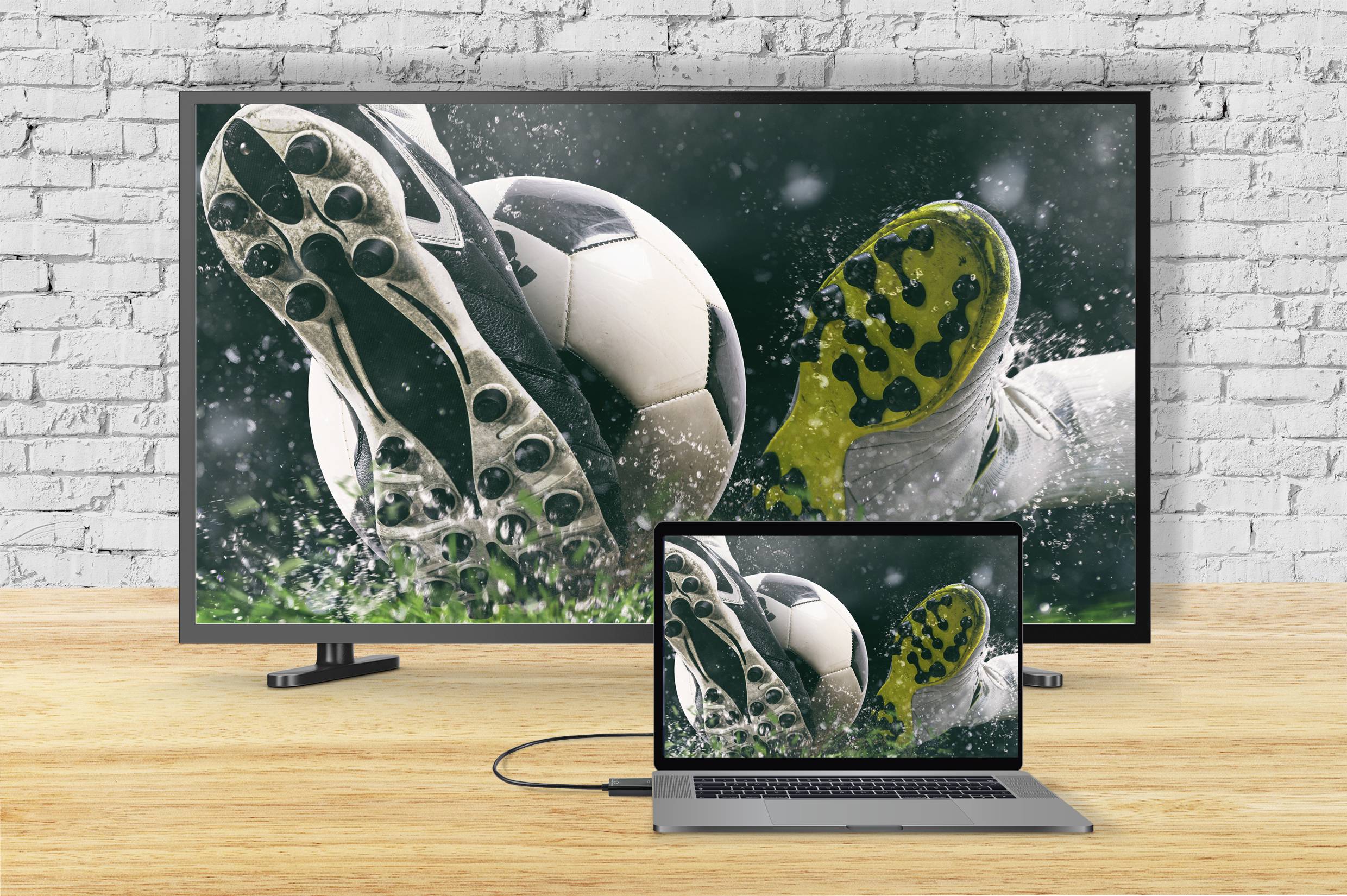 Ein Fernseher und ein Laptop zeigen dasselbe Bild: Fußballschuhe in Aktion, die einen nassen Ball treten.