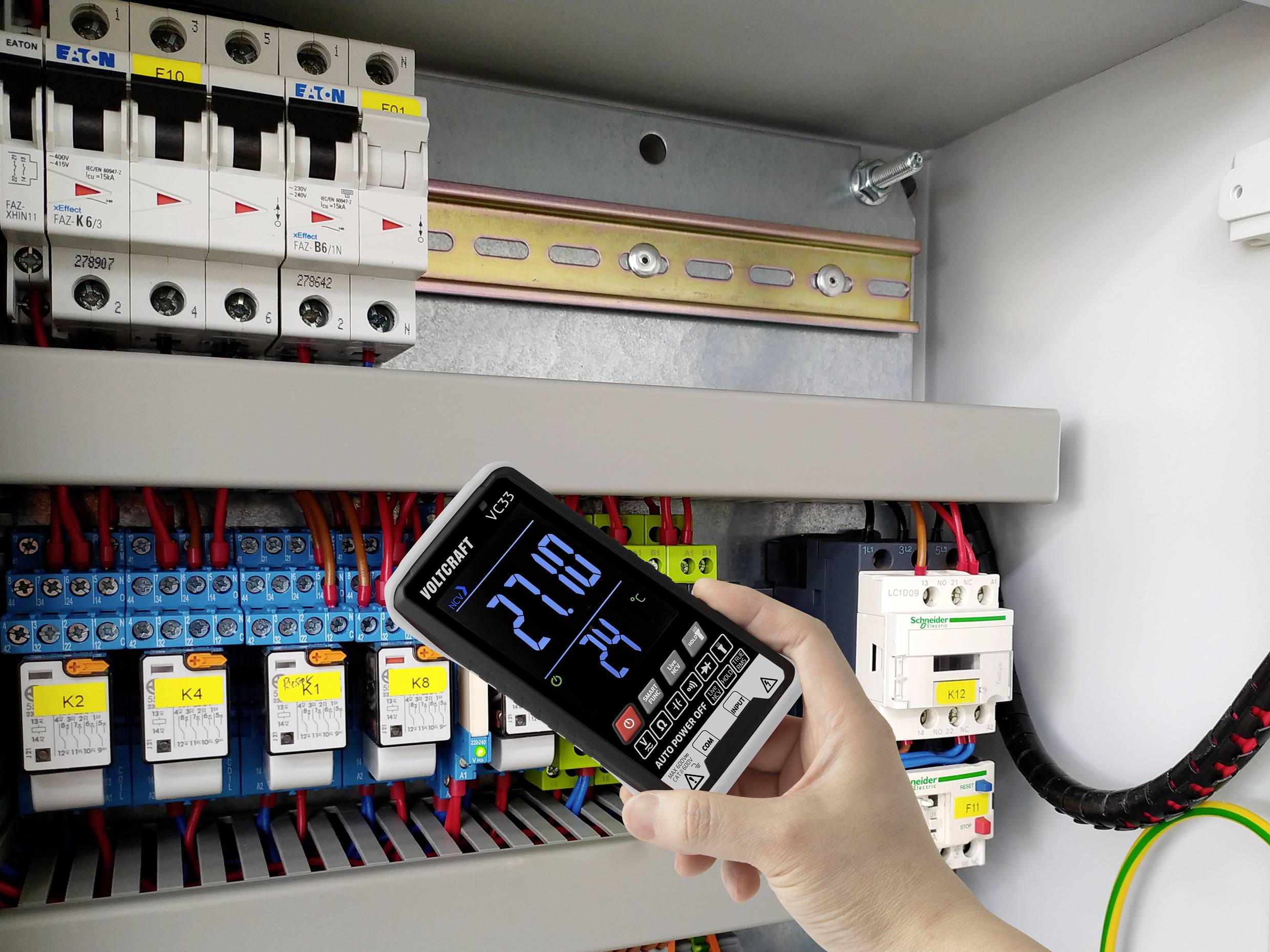 Eine Hand hält ein digitales Messgerät, das '27.10' anzeigt, vor einer geöffneten Schalttafel mit mehreren farbigen Kabeln und Sicherungen.