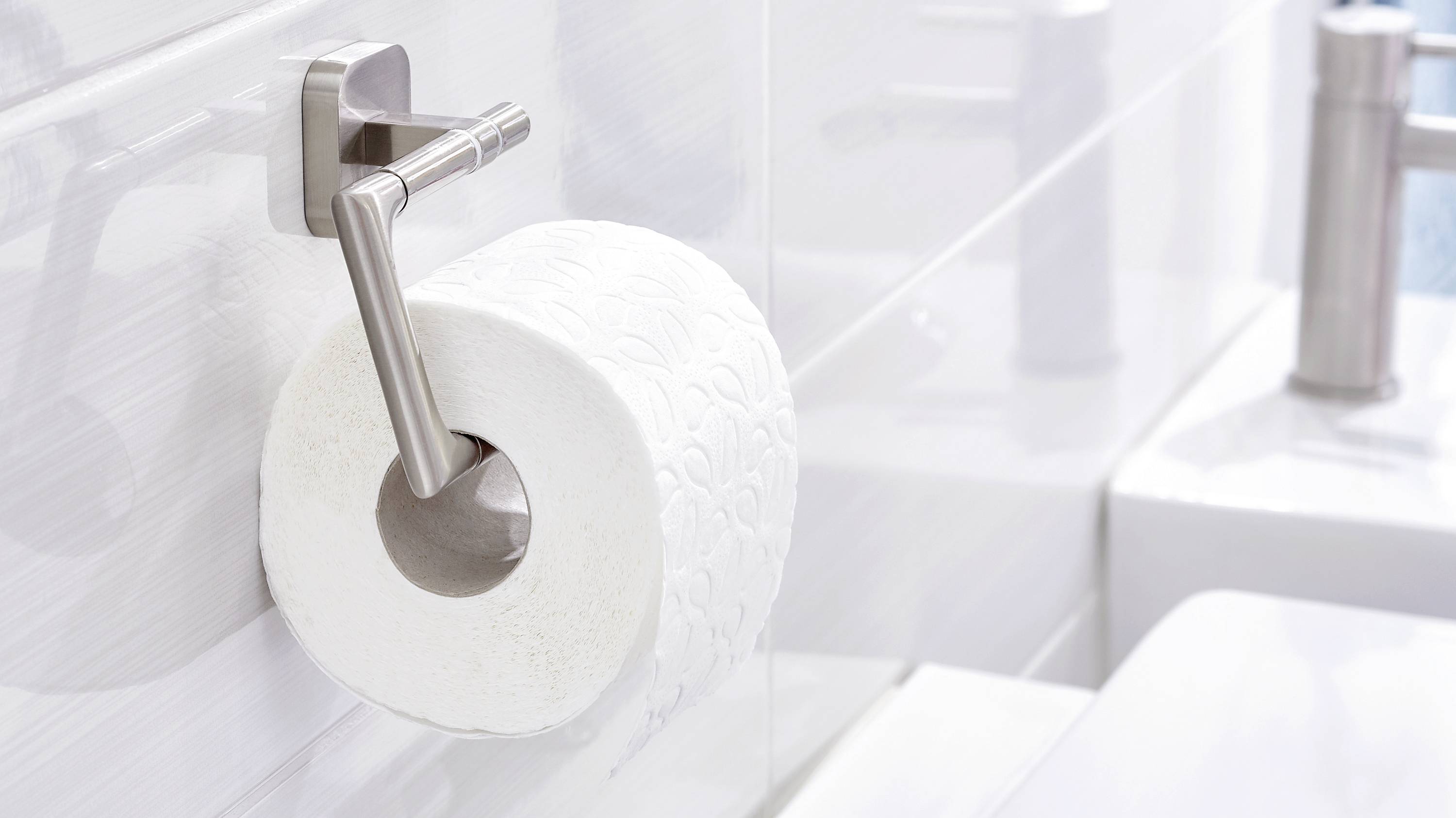 Toilettenpapierrolle auf einem Halter an einer weißen Badezimmerwand, neben einem Waschbecken.