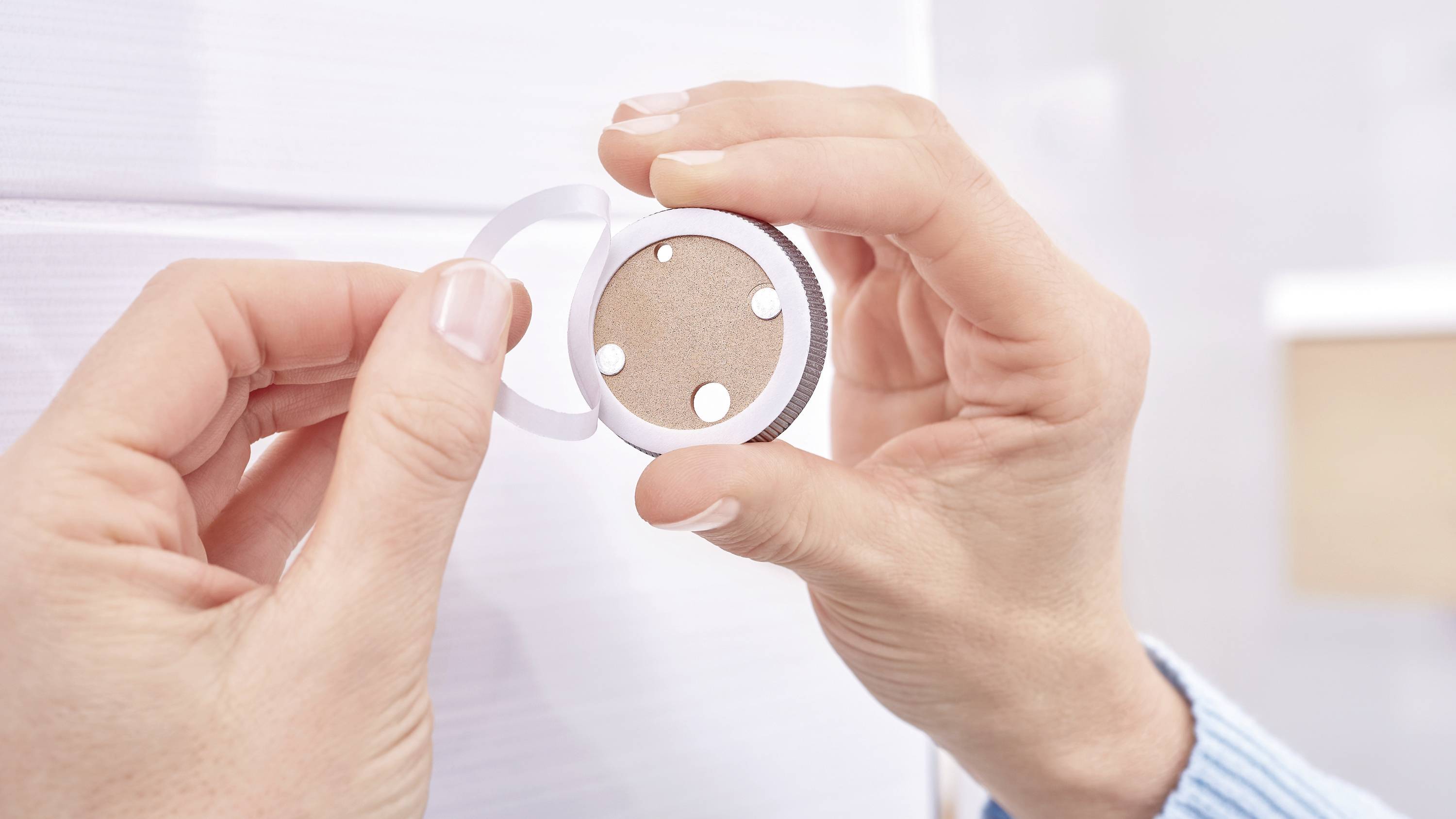 Eine Person hält und installiert ein kleines, rundes, batteriebetriebenes Nachtlicht mit Klebefläche auf einer hellen Wandfläche.