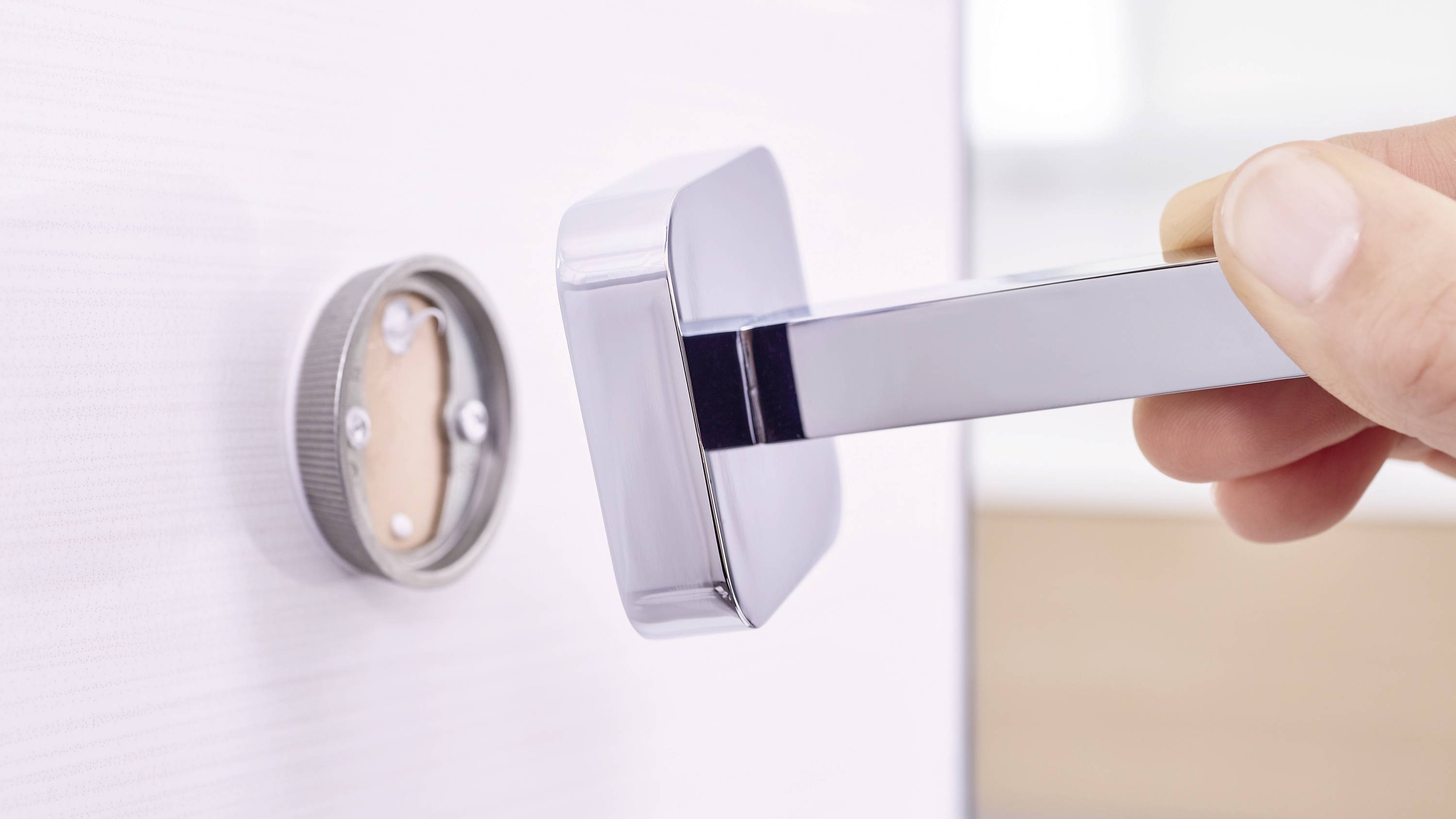 Hand dreht modernen Türgriff an einer weißen Holztür. Fokus auf dem glänzenden metallischen Griff und dem Schließmechanismus.