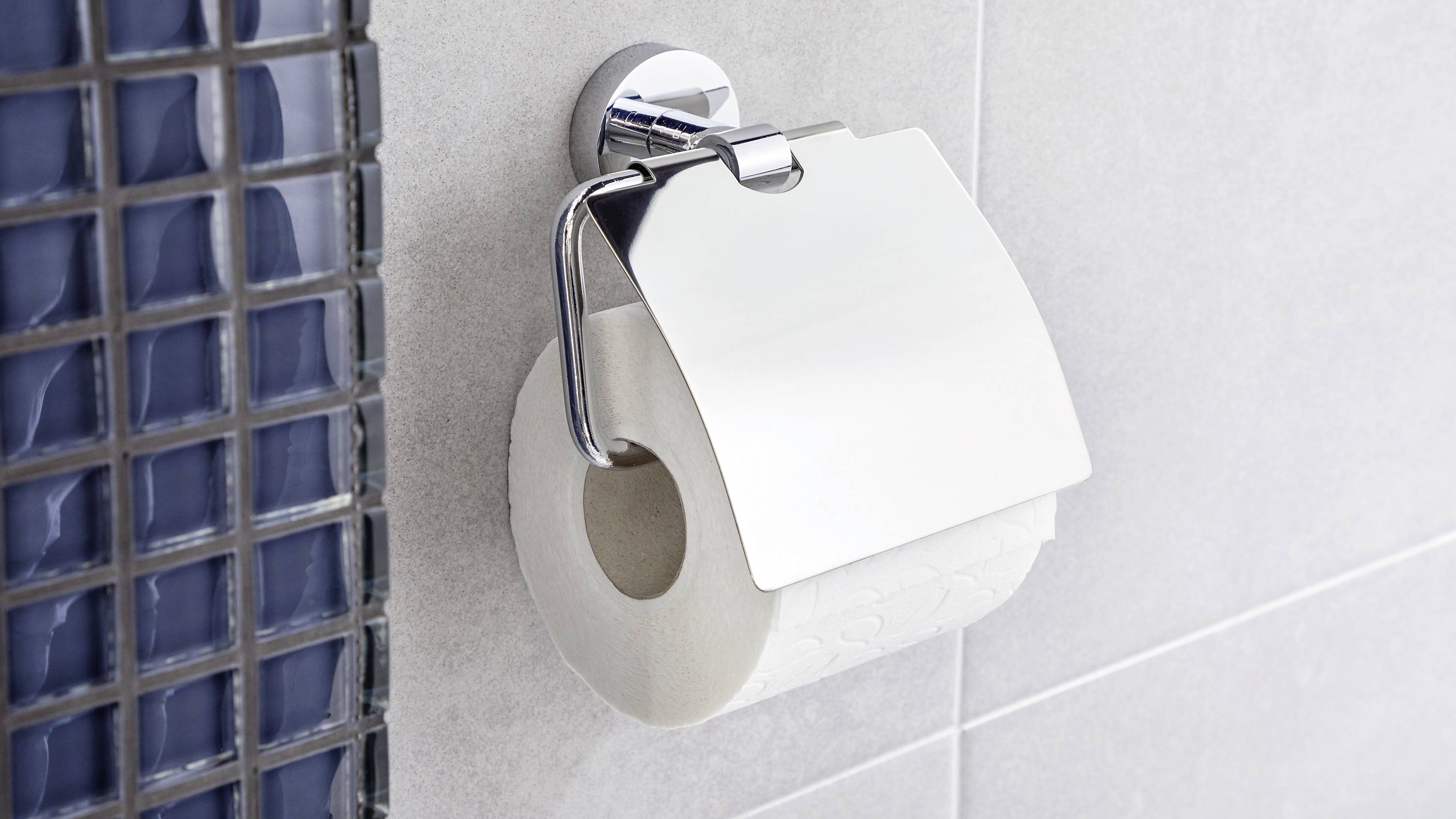Toilettenpapierrolle an einem Halter an der Badezimmerwand, teilweise von einer metallischen Abdeckung bedeckt. Auf hellgrauen Fliesen montiert.