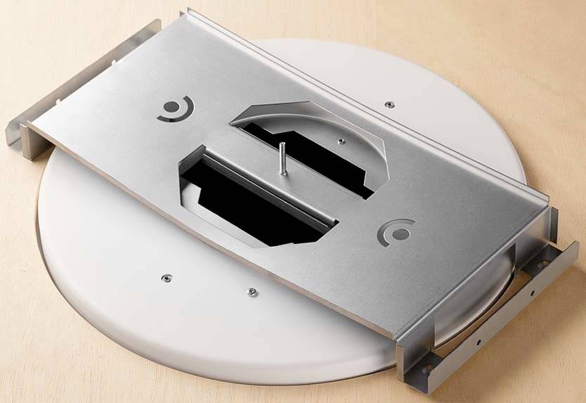 Metallplatte in rechteckiger Form mit zwei Schlitzen auf einer runden weißen Unterlage liegend, Schrauben an den Ecken.