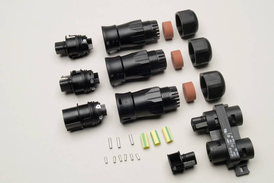 Verschiedene schwarze Stecker und Verbindungsteile auf hellgrauem Hintergrund, geordnet in Reihen mit metallischen und farbigen Einsätzen.