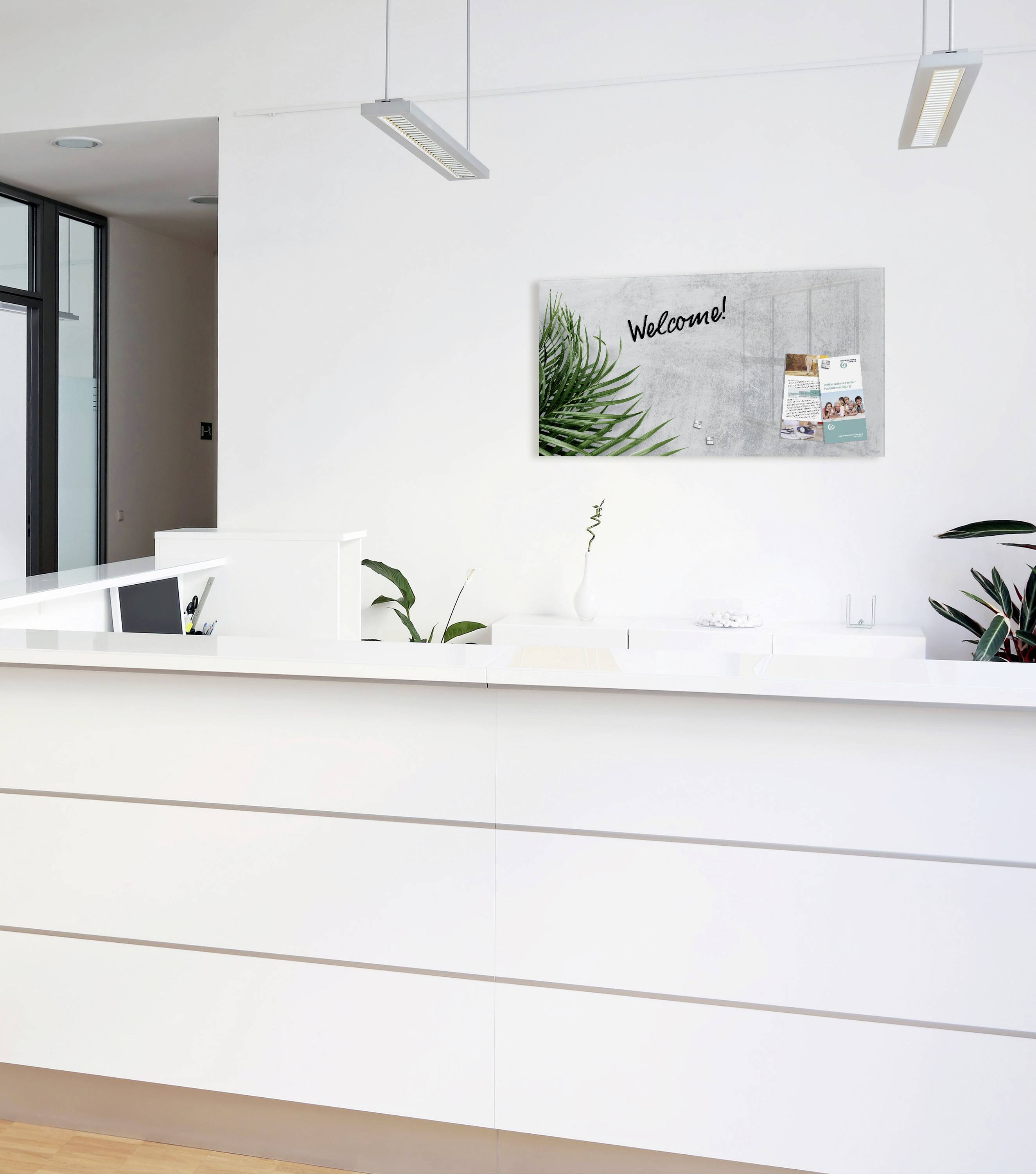 Empfangsbereich mit modernem, weißem Tresen, darüber ein Bild mit 'Welcome!', Pflanzen und Broschüren an der Wand.