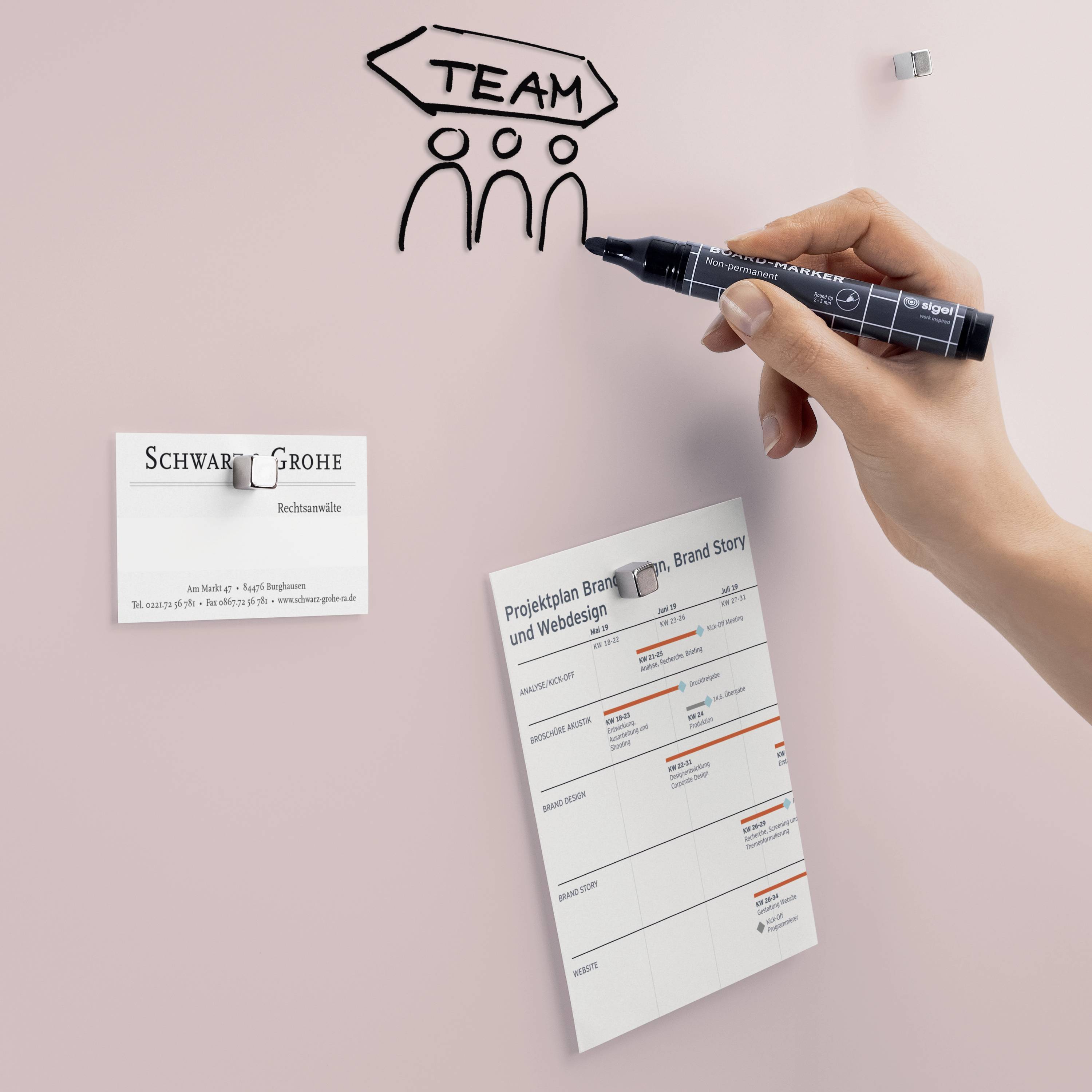 'Team' wird mit einem schwarzen Stift auf eine rosa Wand gemalt. Nebenstehend sind eine Visitenkarte und ein Projektplaner angebracht.