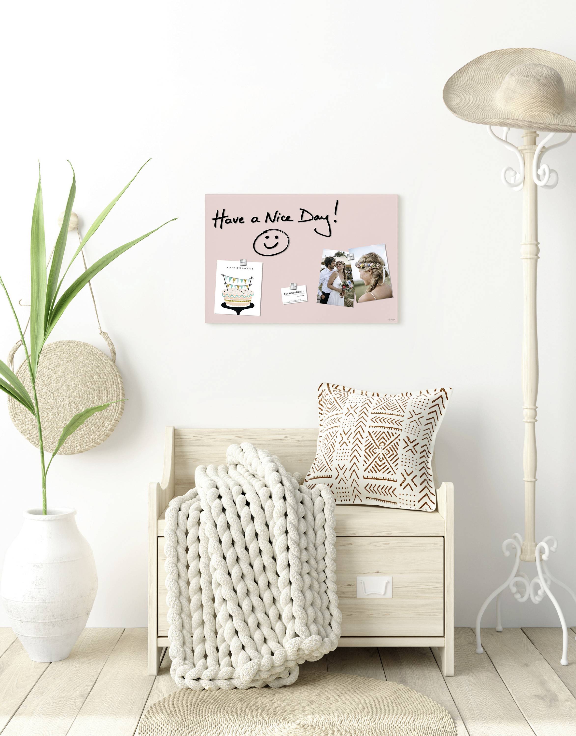 Ein heller Raum mit einem Holzbänkchen, einer Pflanze und einem großen Hut. An der Wand hängt ein Schild mit der Aufschrift 'Have a Nice Day!'.