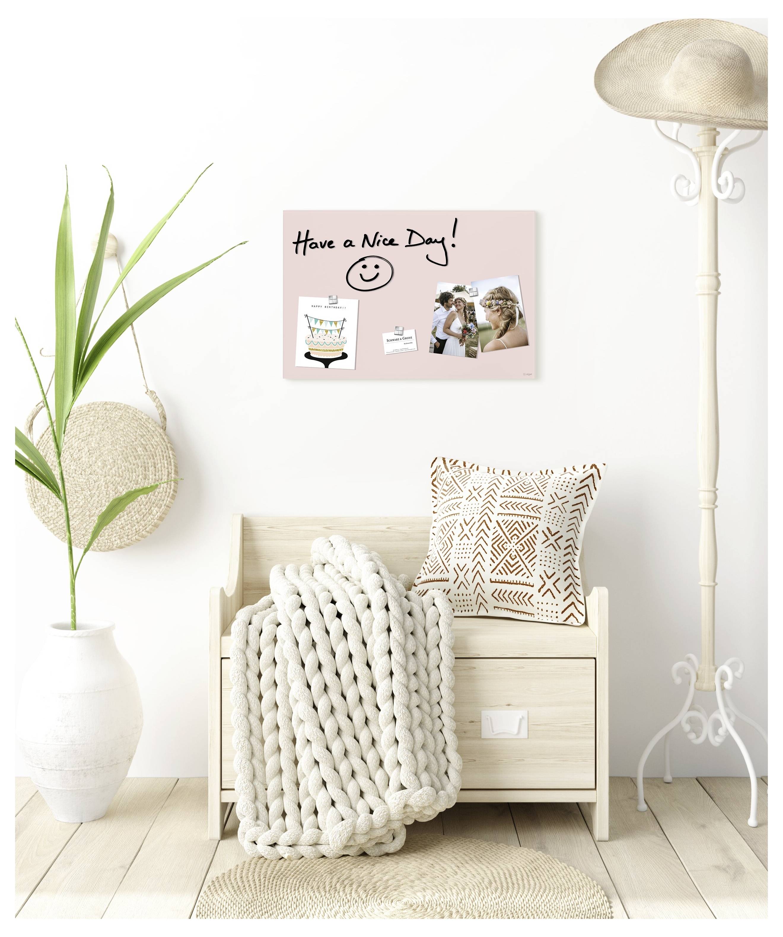 Helles minimalistisches Zimmer mit einer Holzbank, die mit einer dicken Strickdecke drapiert ist, einem gemusterten Kissen, einer Pflanze, einer Stehlampe und einem Wandschild mit der Aufschrift „Have a Nice Day!"