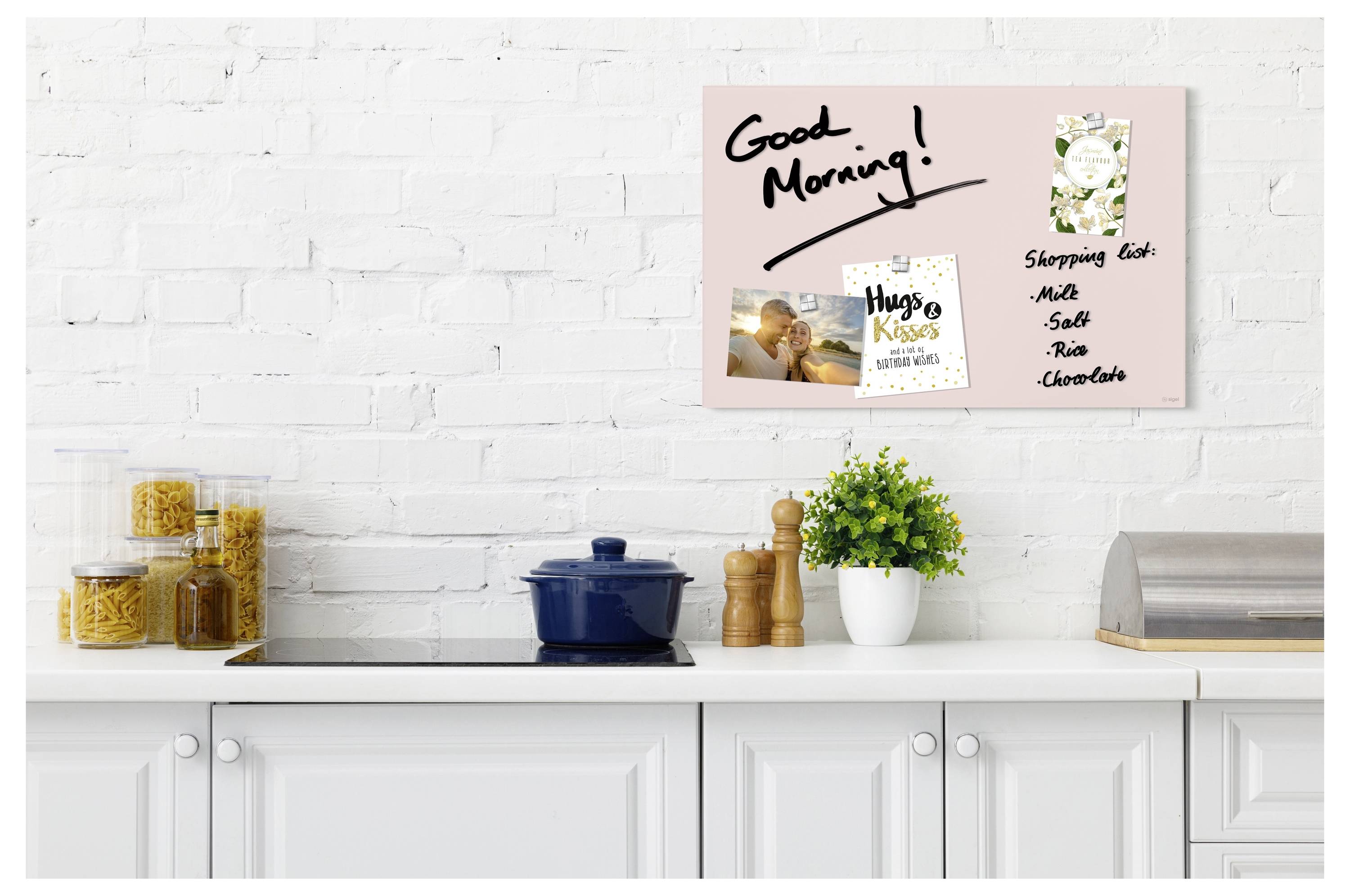 Küchentheke mit einem rosa Notizbrett mit der Aufschrift "Guten Morgen!" und einer Einkaufsliste, über weißen Schränken und Arbeitsplattendekorationen.