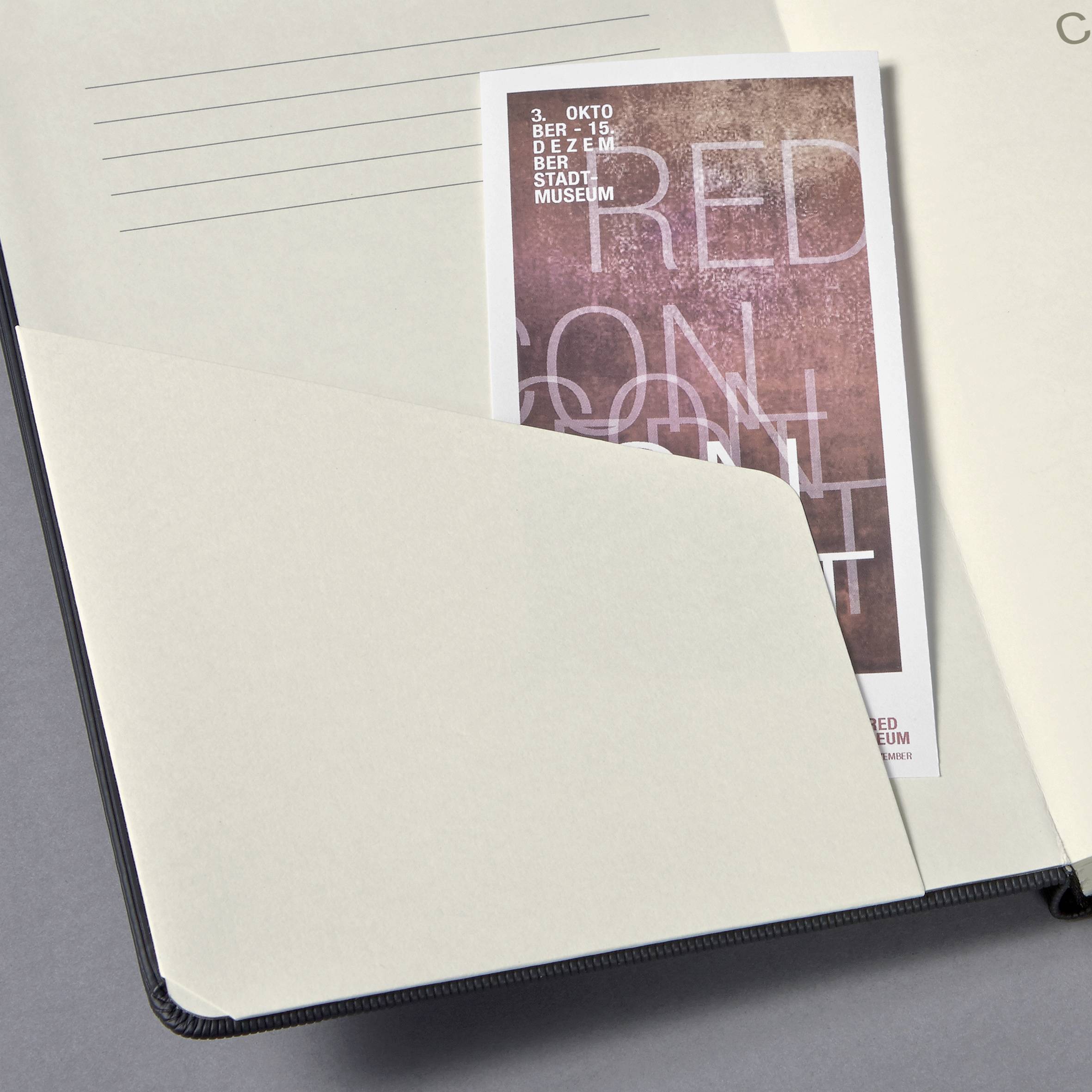 Eine Postkarte mit Text: '3. Oktober, der 25. Stadtmuseum'. Sie liegt schräg in einem Notizbuch mit linierten Seiten.