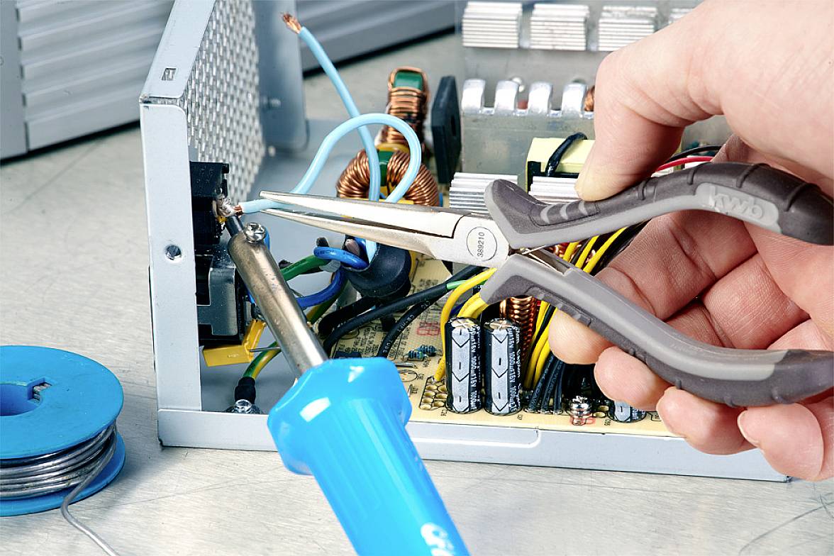 Eine Hand hält eine Zange bei der Reparatur eines elektronischen Geräts. Ein Lötkolben und Drähte sind sichtbar.