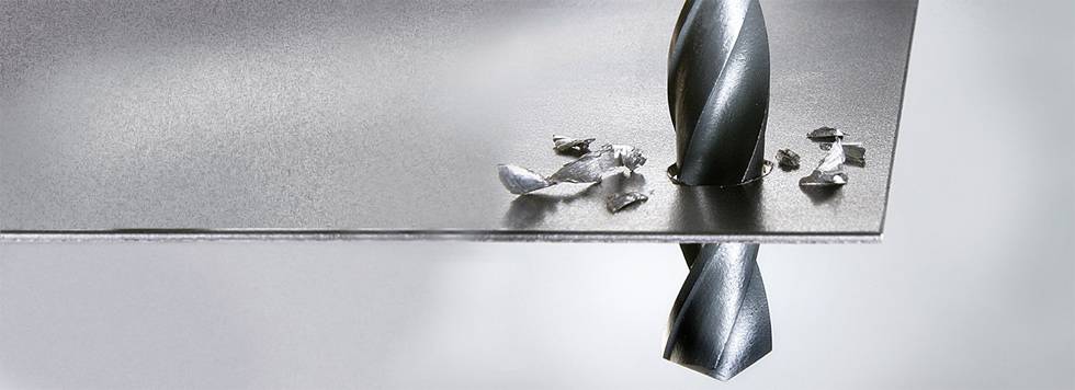 Ein Metallbohrer durchbohrt ein dünnes Metallblech, während Späne um das gebohrte Loch herum verstreut sind.