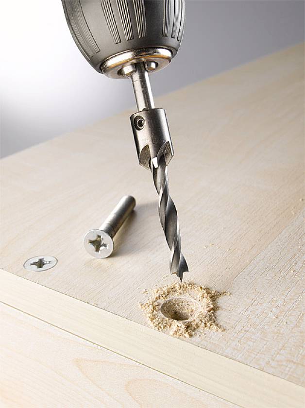 Bohrer mit Holzbohrer, bohrt ein Loch in ein Holzbrett. Zwei Schrauben liegen daneben.