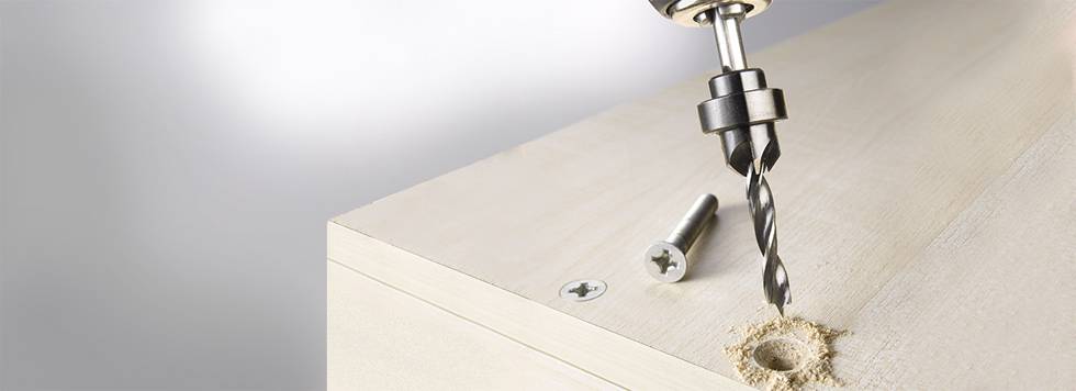Bohrer erstellt Loch in Holzplatte, daneben eine Schraube.