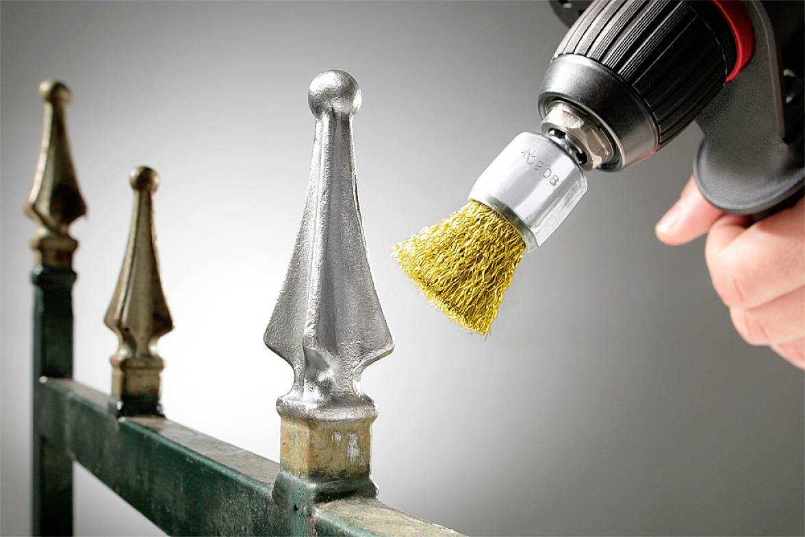 Eine Person schleift mit einer Drahtbürste an einer Bohrmaschine Rost von einem Metallgeländer ab, um es zu reinigen und aufzufrischen.