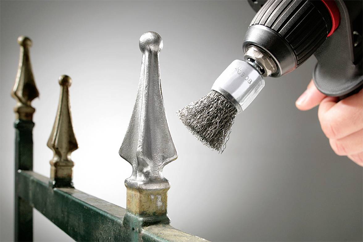 Eine Hand benutzt eine Bohrmaschine mit Drahtbürstenaufsatz, um Rost von einem metallenen Zaunpfosten zu entfernen.