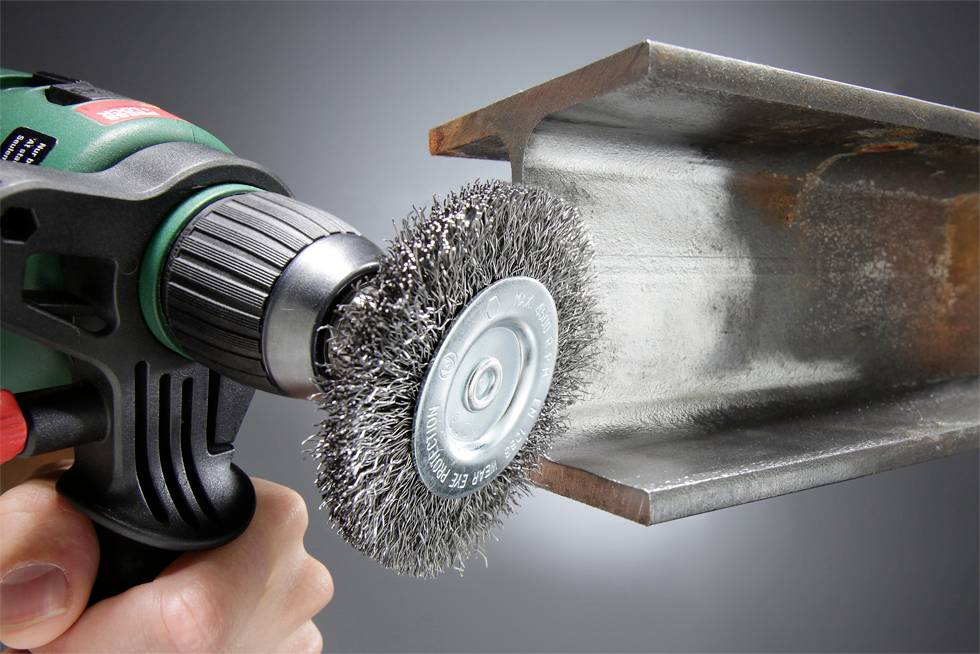 Eine Hand verwendet eine Bohrmaschine mit einem Drahtbürstenaufsatz, um einen Stahlträger zu reinigen und Rost sowie Ablagerungen zu entfernen.