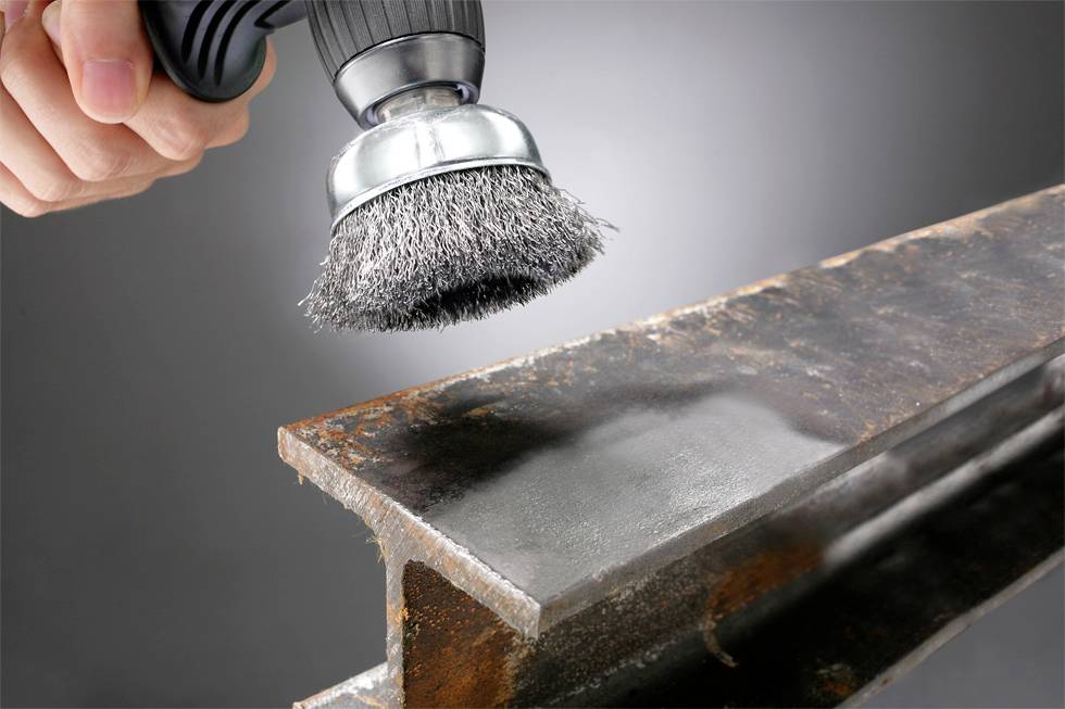 Eine Hand hält eine Drahtbürste und poliert einen rostigen Metallträger, wobei eine glatte, glänzende Oberfläche zum Vorschein kommt. Der Hintergrund ist in neutralem Grau gehalten.