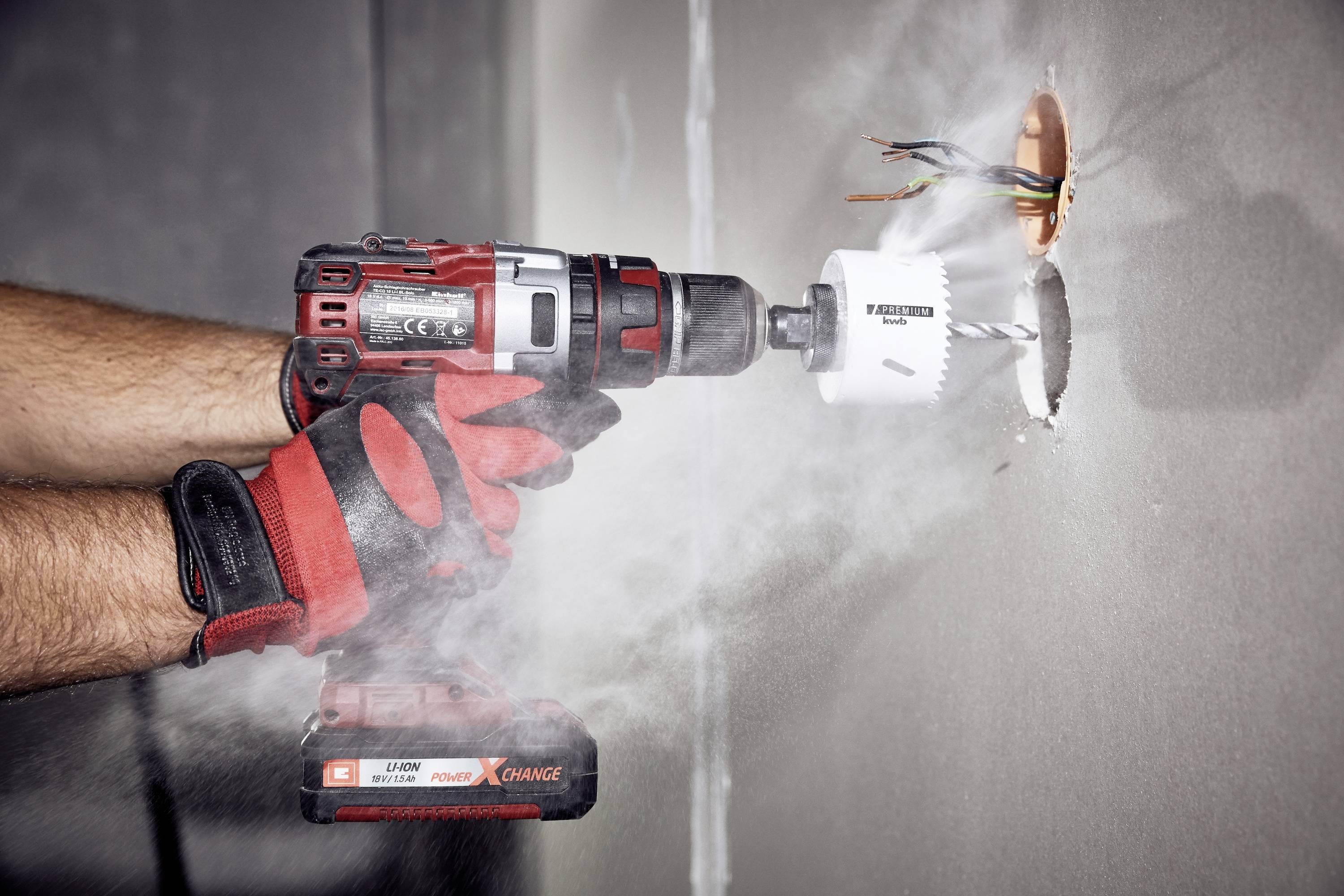 Person trägt Handschuhe und bohrt mit einer elektrischen Bohrmaschine ein Loch in die Wand. Staub wirbelt um die Bohrstelle auf.