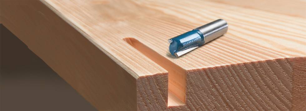 Ein Holzbrett mit einer gefrästen Nut, daneben ein metallener Fräser mit blauem Einsatz, platziert auf dem Tisch.