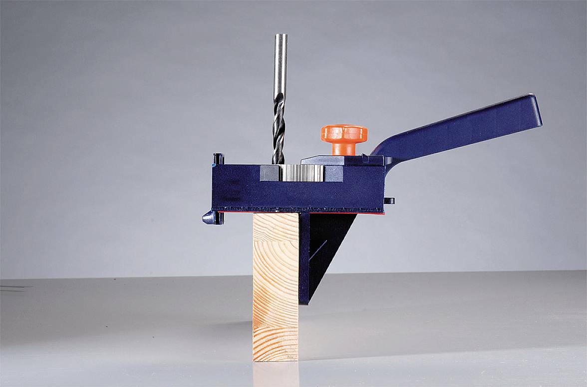 Ein Bohrer in einer blauen Halterung bohrt ein Loch in einen Holzblock auf einem Tisch. Ein orangefarbener Griff ist sichtbar.