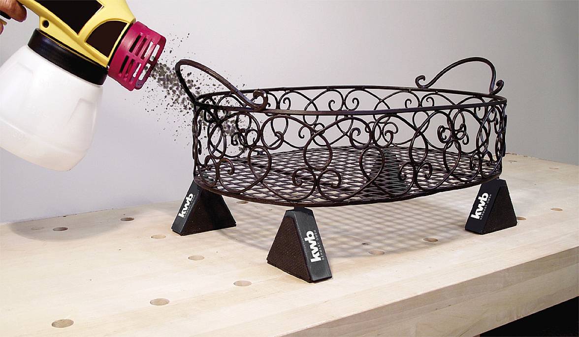 Ein Metallkorb wird auf einem Tisch mit einer Sprühpistole schwarz besprüht. Kleine, schwarze Lacktröpfchen verteilen sich gleichmäßig.