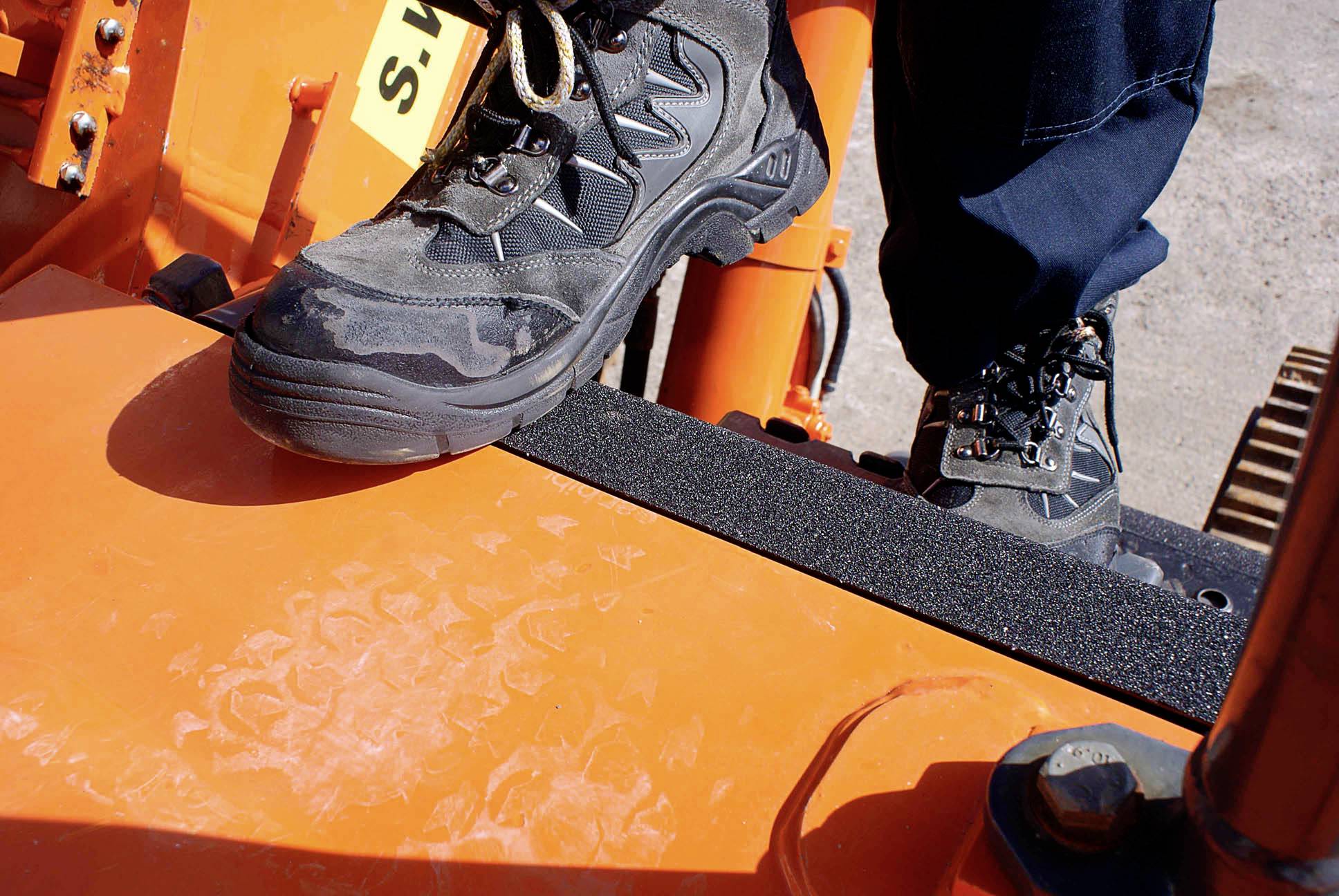 Ein Arbeiter steht mit robusten Wanderschuhen auf einer orangefarbenen Plattform einer schweren Baumaschine.