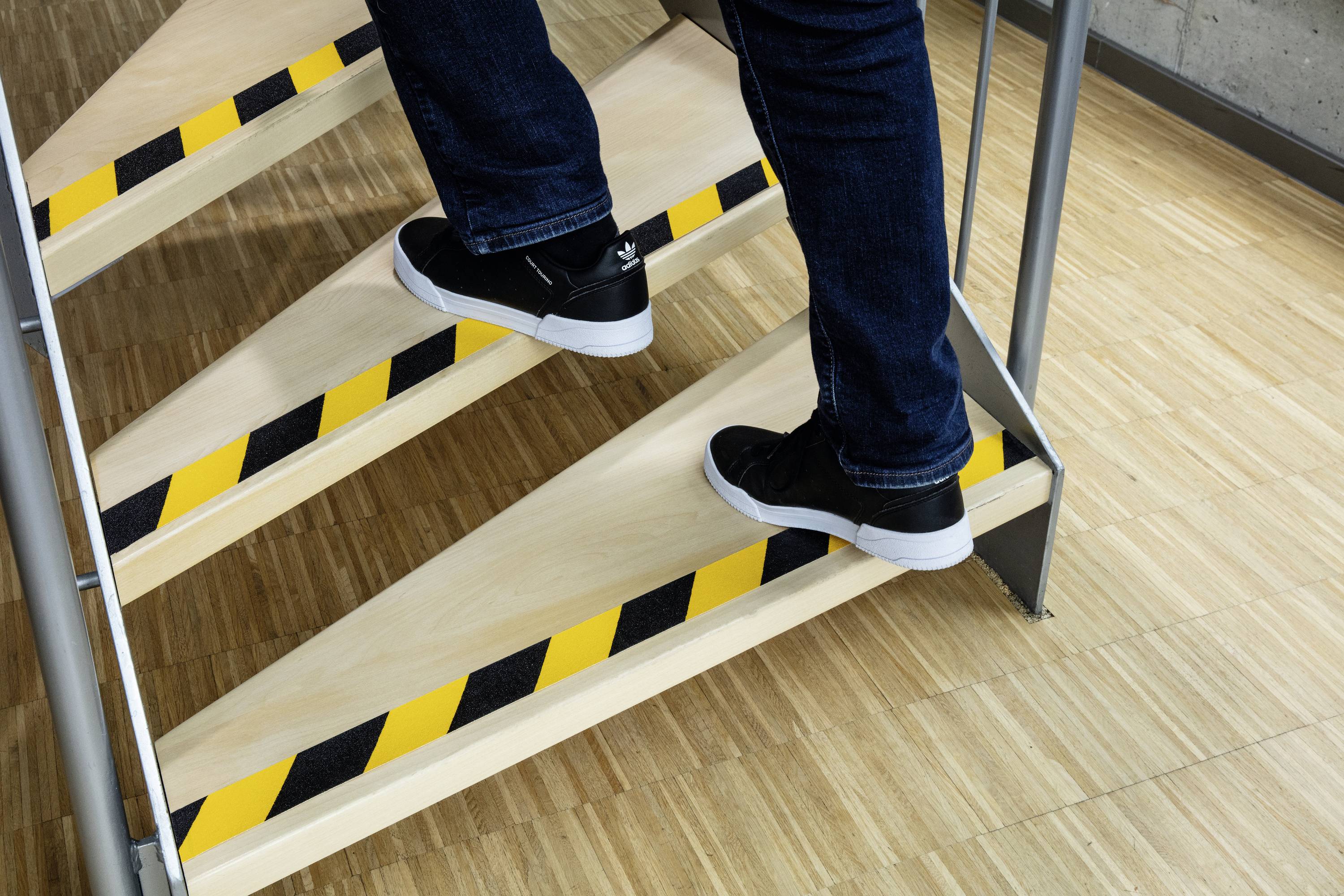 Nahaufnahme von jemandem, der eine Treppe mit gelb-schwarzen Warnmarkierungen an den Stufen hinaufsteigt, getragen werden Jeans und schwarze Turnschuhe.